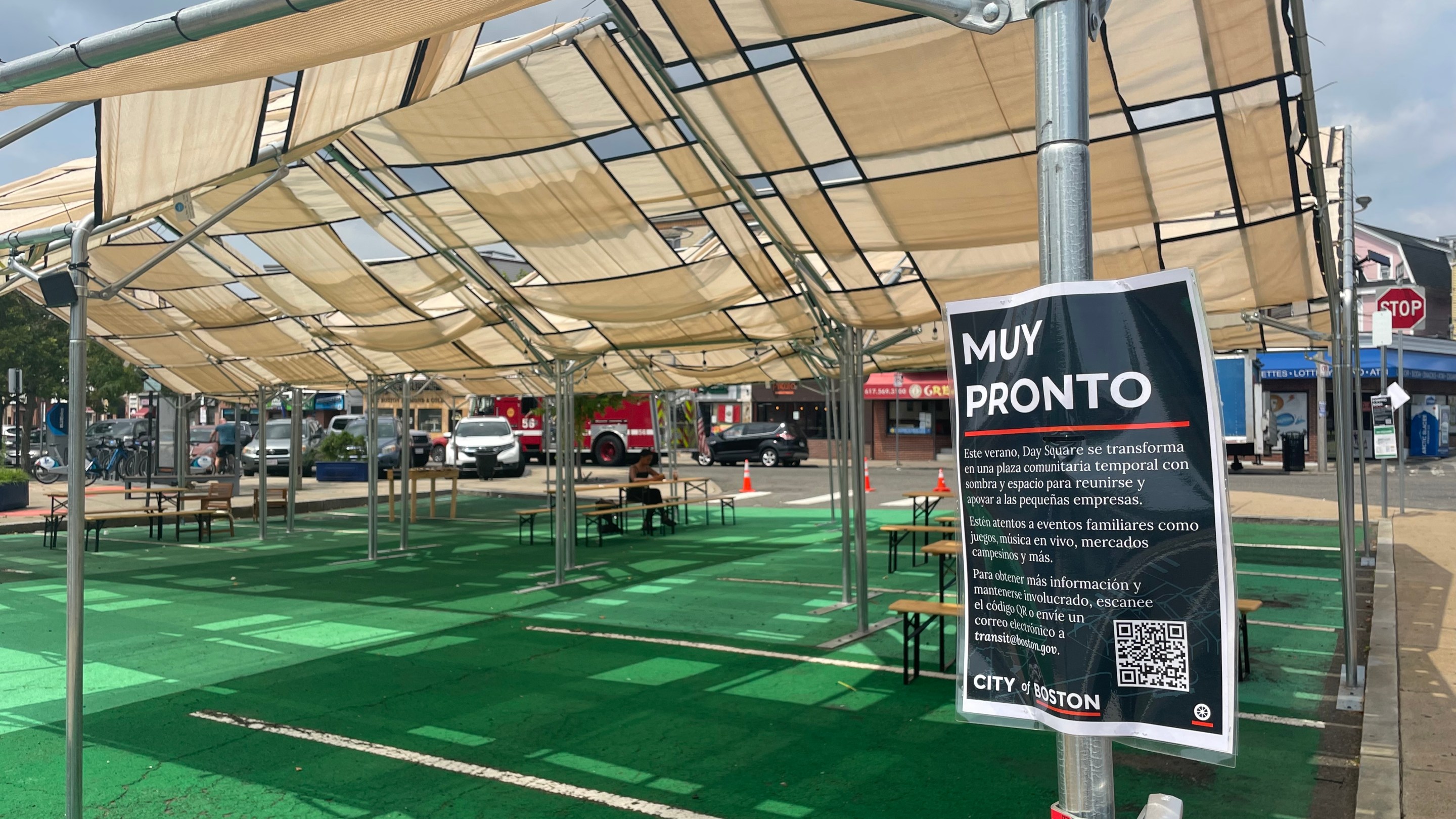 A shade structure over green-painted pavement. A spanish language sign in the foreground, attached to a tent post, is titled "Muy Pronto" (coming soon) and explains that the temporary plaza will host "eventos familiares como juegos, música en vivo, mercados campesinos y más"