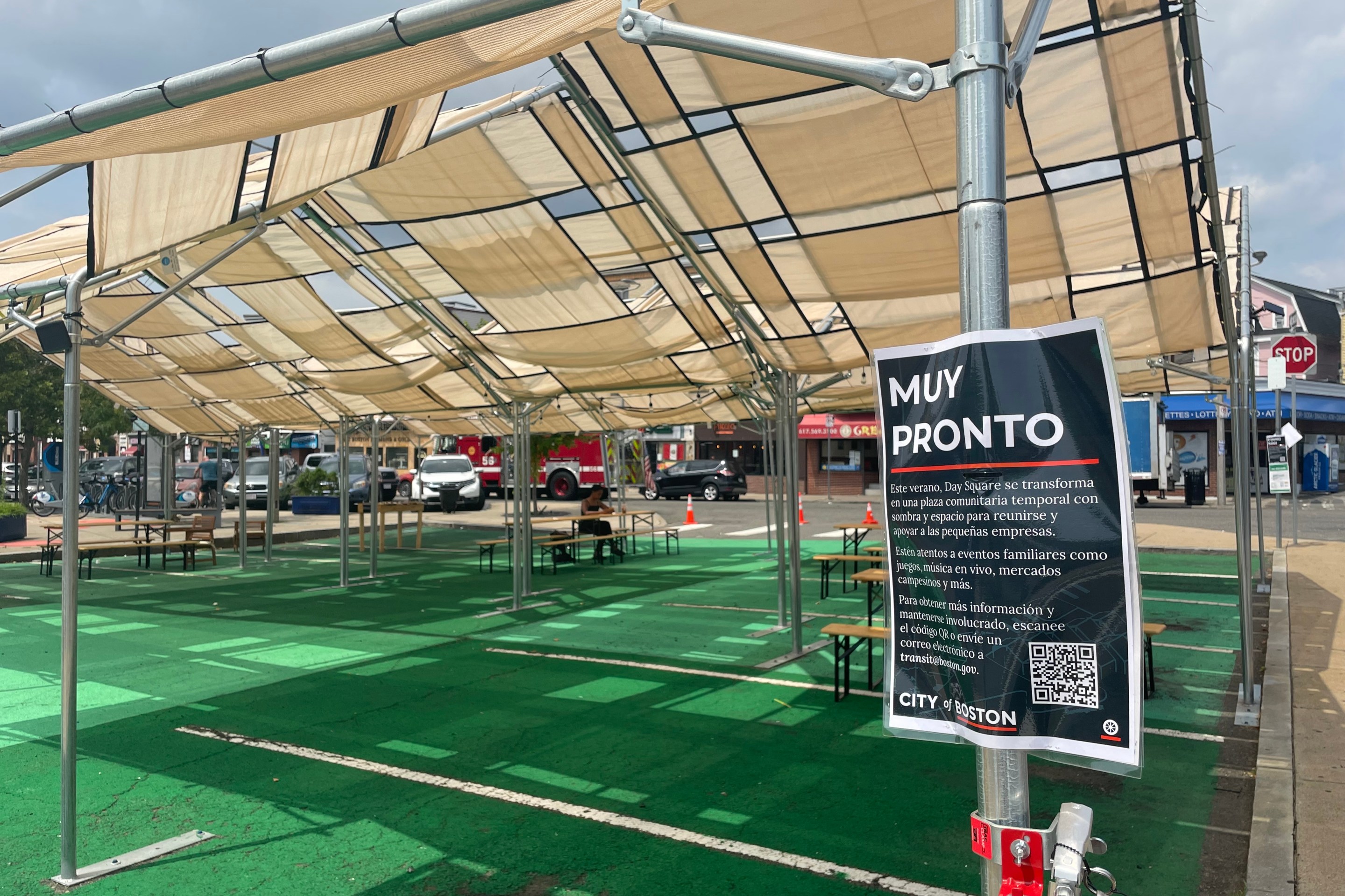 A shade structure over green-painted pavement. A spanish language sign in the foreground, attached to a tent post, is titled "Muy Pronto" (coming soon) and explains that the temporary plaza will host "eventos familiares como juegos, música en vivo, mercados campesinos y más"