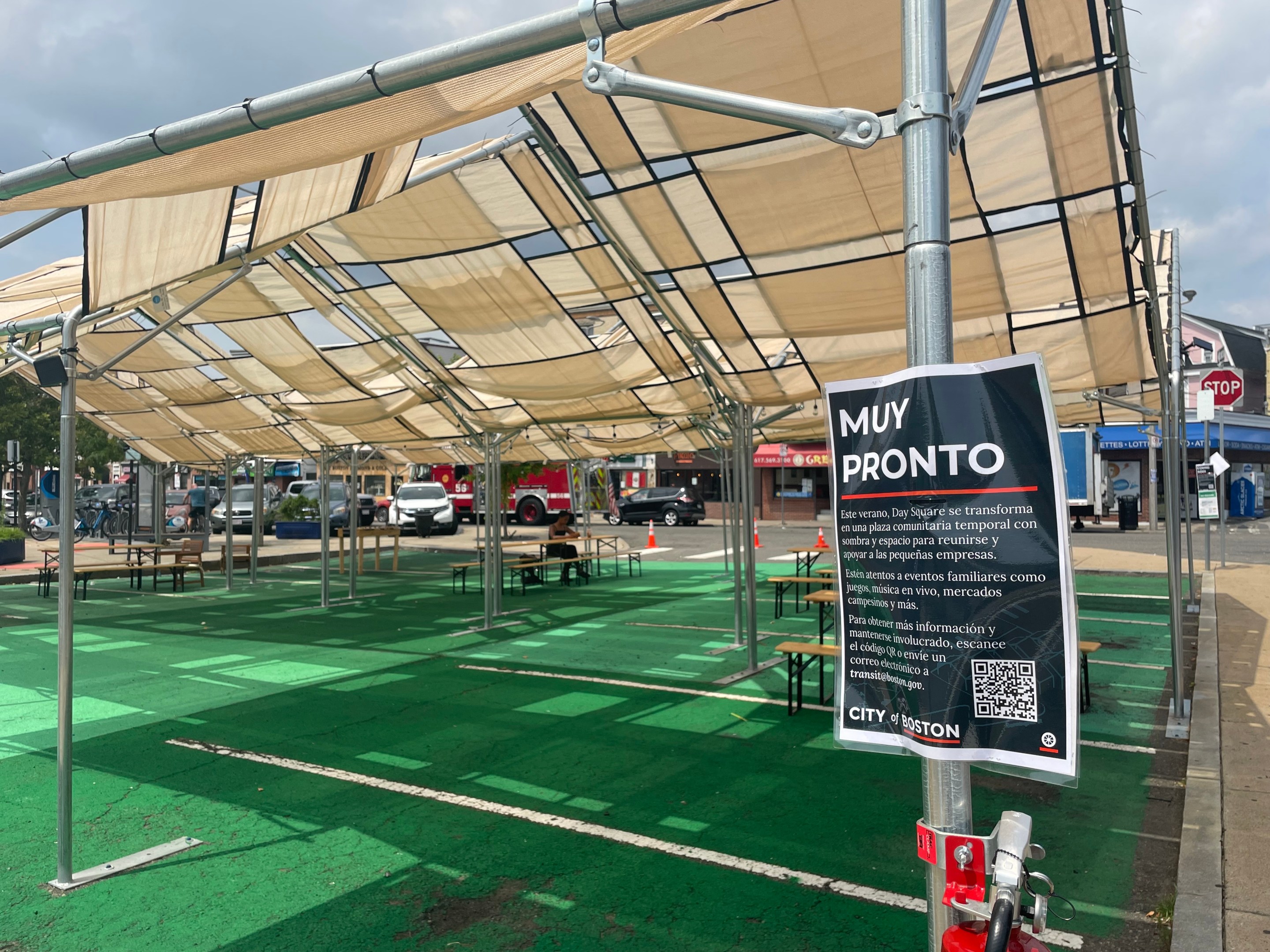 A shade structure over green-painted pavement. A spanish language sign in the foreground, attached to a tent post, is titled "Muy Pronto" (coming soon) and explains that the temporary plaza will host "eventos familiares como juegos, música en vivo, mercados campesinos y más"