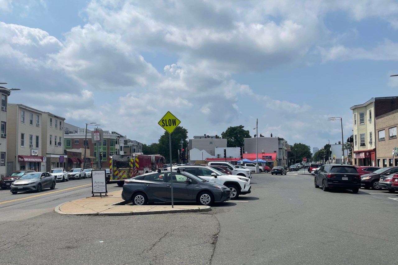 A large parking lot next to a two-lane city street, surrounded by 3- and 4-story buildings. In the foreground is a diamond-shaped "SLOW" sign