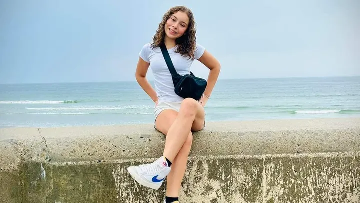 A smiling teen girl sits on top of a wall near the ocean.