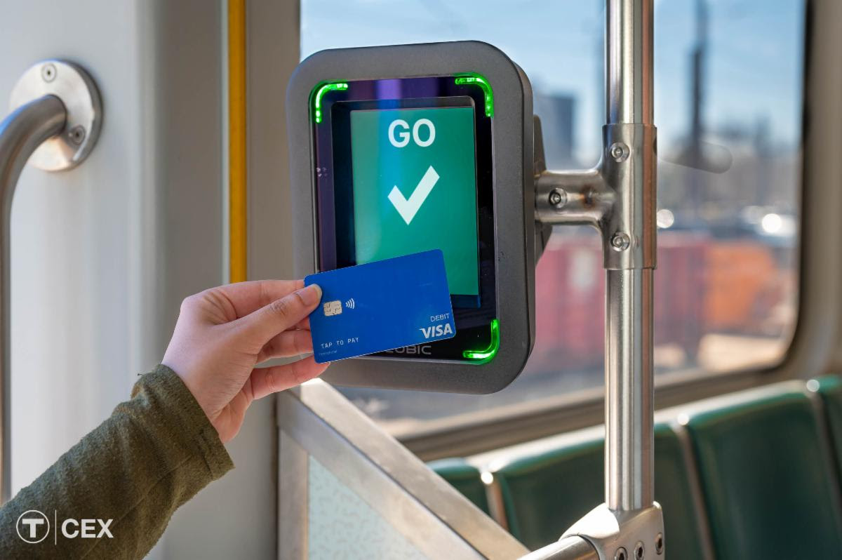 A person holds a credit card against a small screen that displays the word "GO" above a checkmark on a green background. The screen is mounted to a pole inside a transit vehicle.
