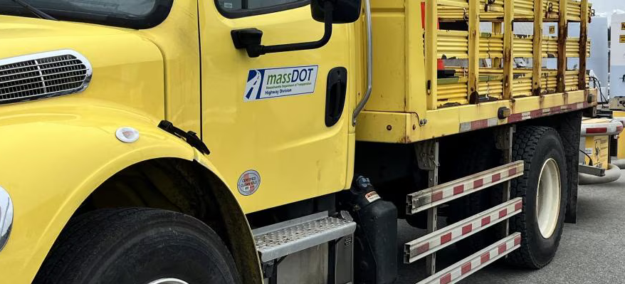 A yellow work truck with a MassDOT logo on the door and lateral protective devices on the undercarriage, marked with red and white stripes, between the wheels.