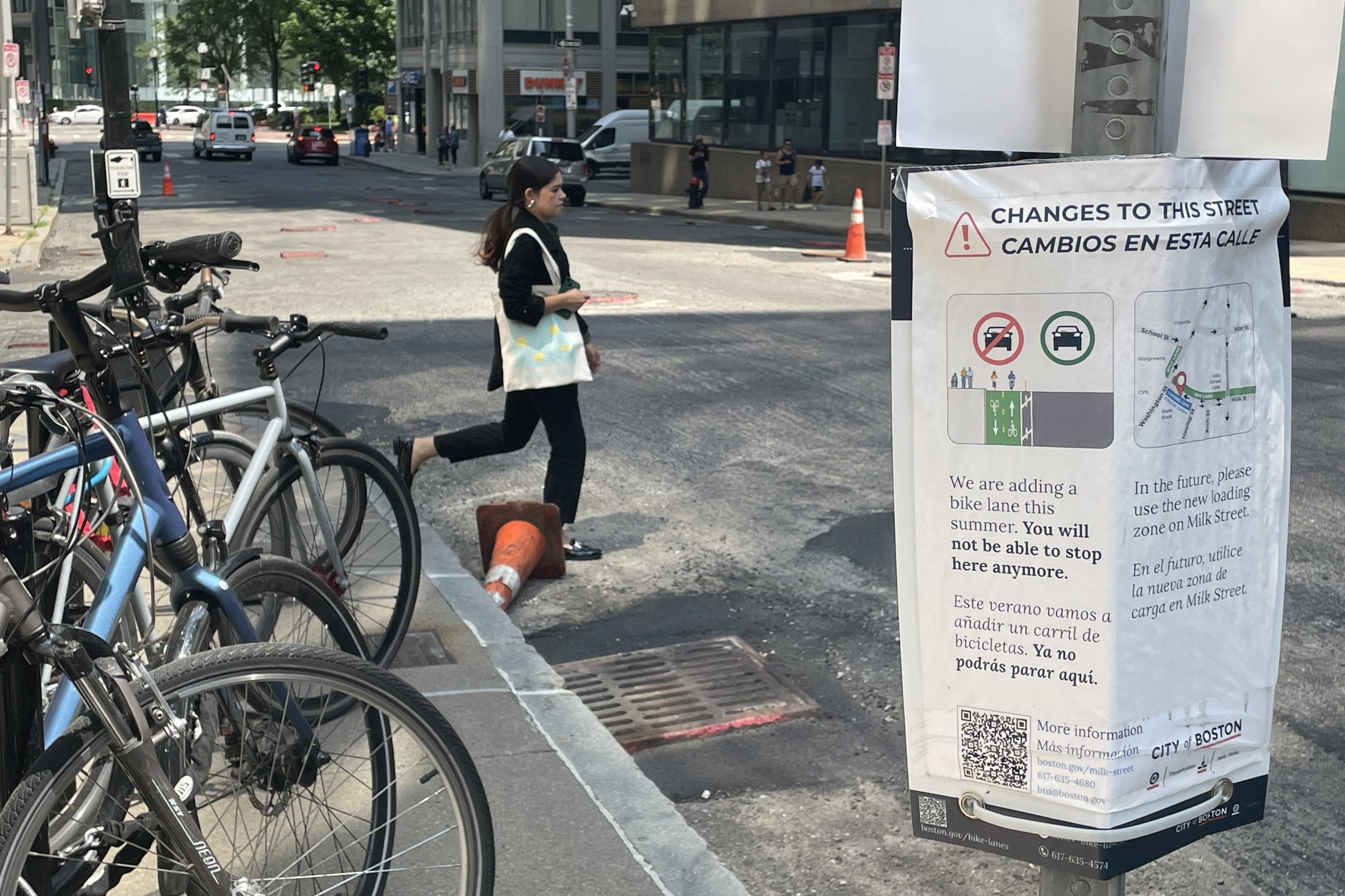A pedestrian steps into a street next to a full bike rack. In the foreground, a flyer attached to a sign post announces an upcoming paving project to add new bike lanes to the street.