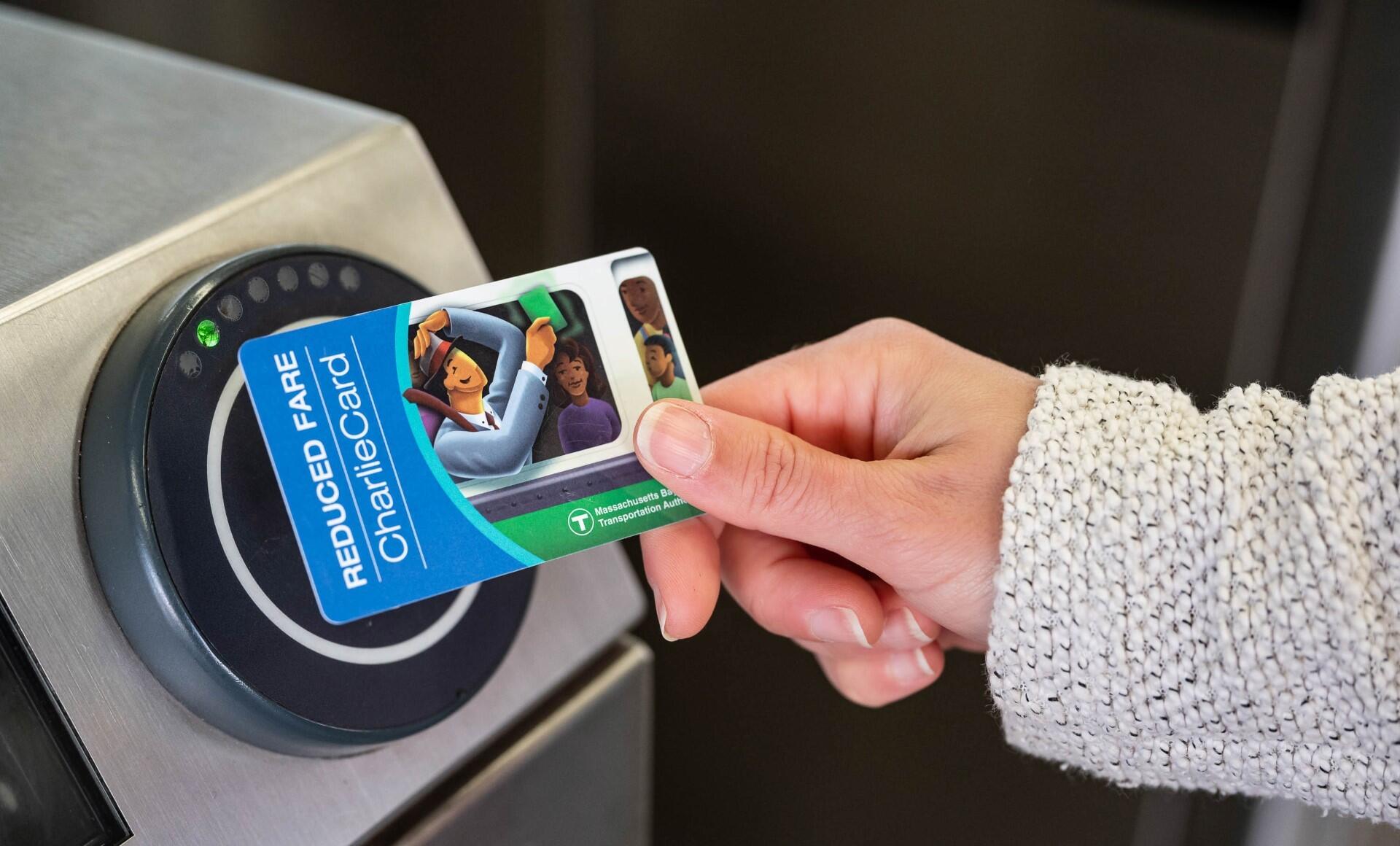 A photo of a hand holding a blue "Reduced Fare CharlieCard" against a fare reader at a subway entrance gate.