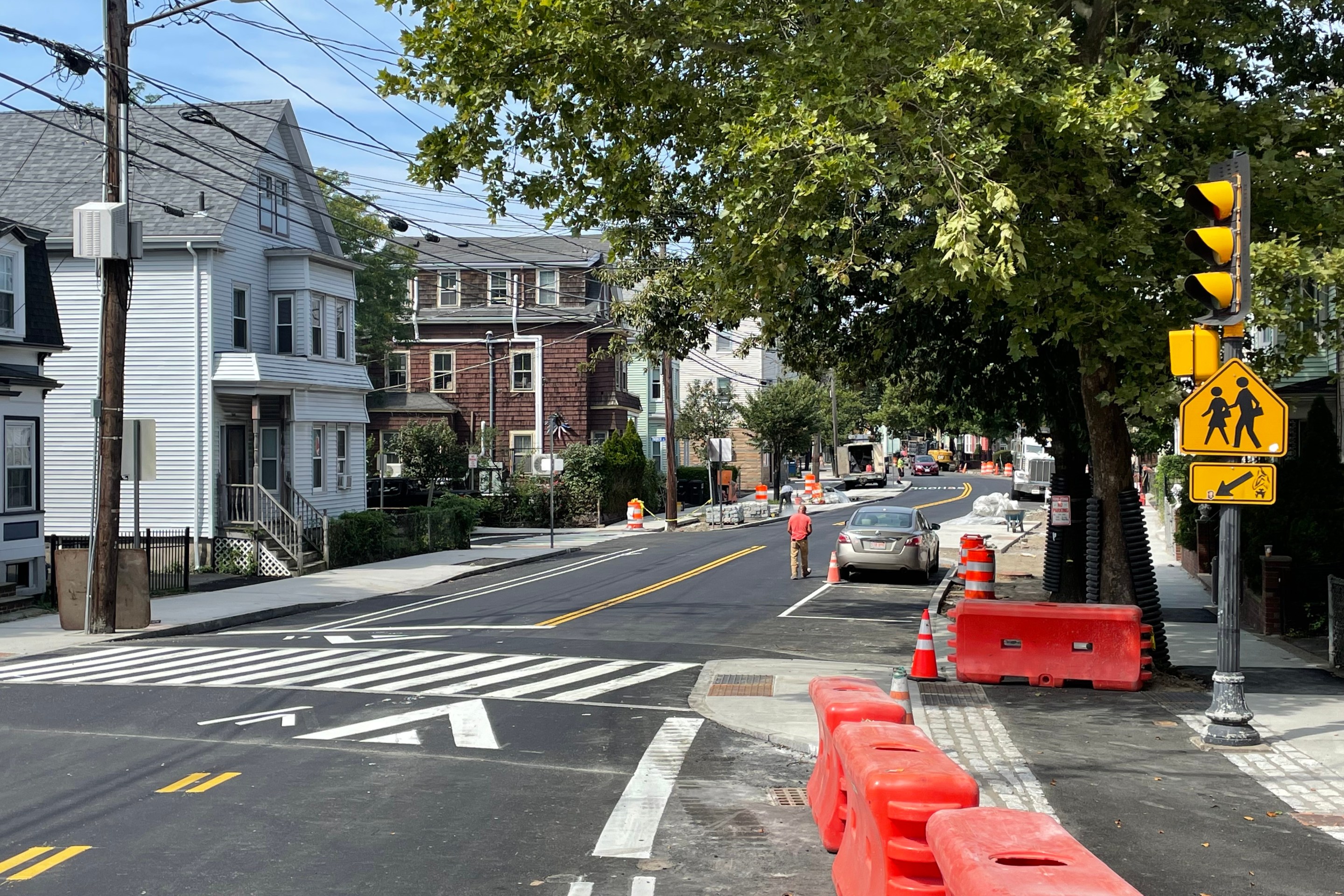 A new sidewalk-level bike lane under construction adjacent to a two-lane city street. Cones and construction barrels litter the bike lane. In the foreground is a raised crosswalk next to a traffic signal.