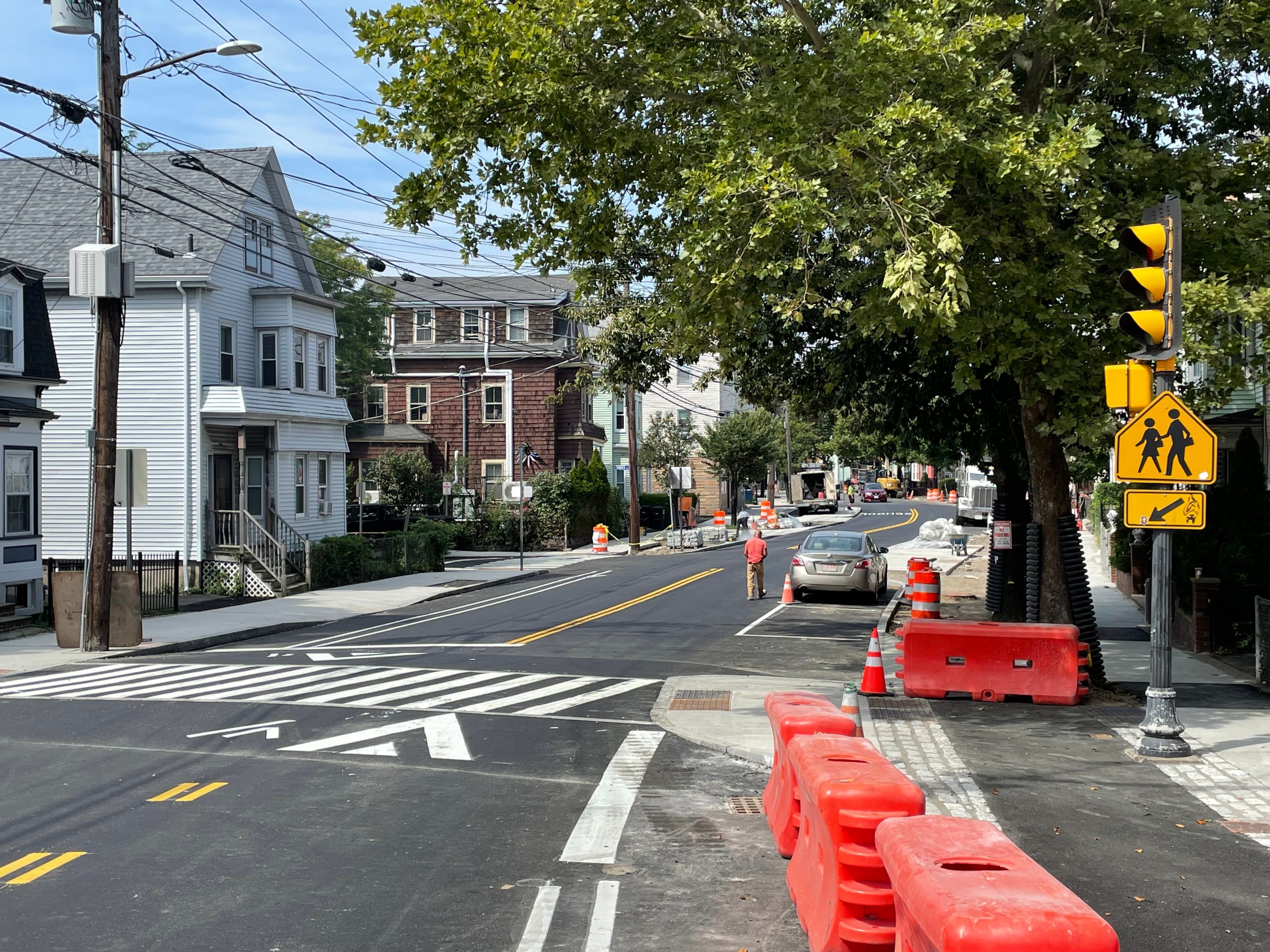 A new sidewalk-level bike lane under construction adjacent to a two-lane city street. Cones and construction barrels litter the bike lane. In the foreground is a raised crosswalk next to a traffic signal.