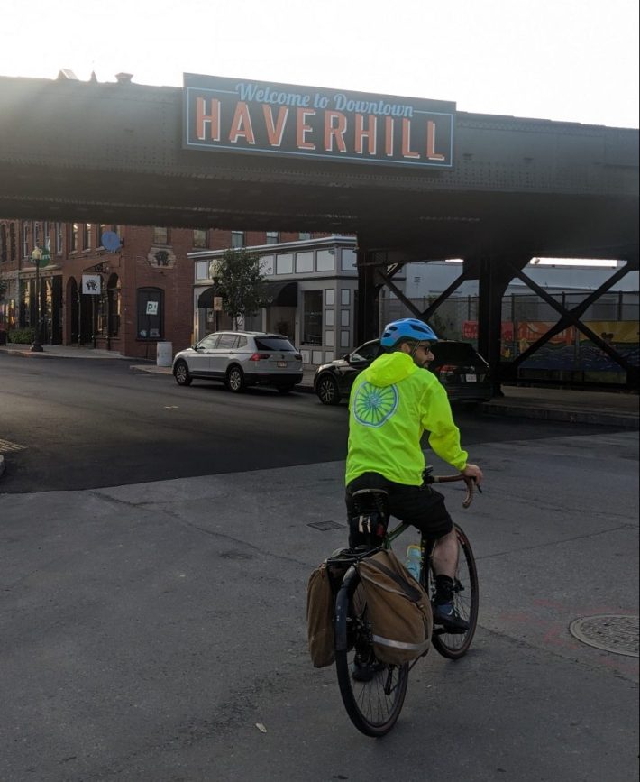 A man on a bike with panniers, wearing a dayglo yellow hooded jacket, rides towards a railroad bridge with a sign that says "welcome to downtown Haverhill"