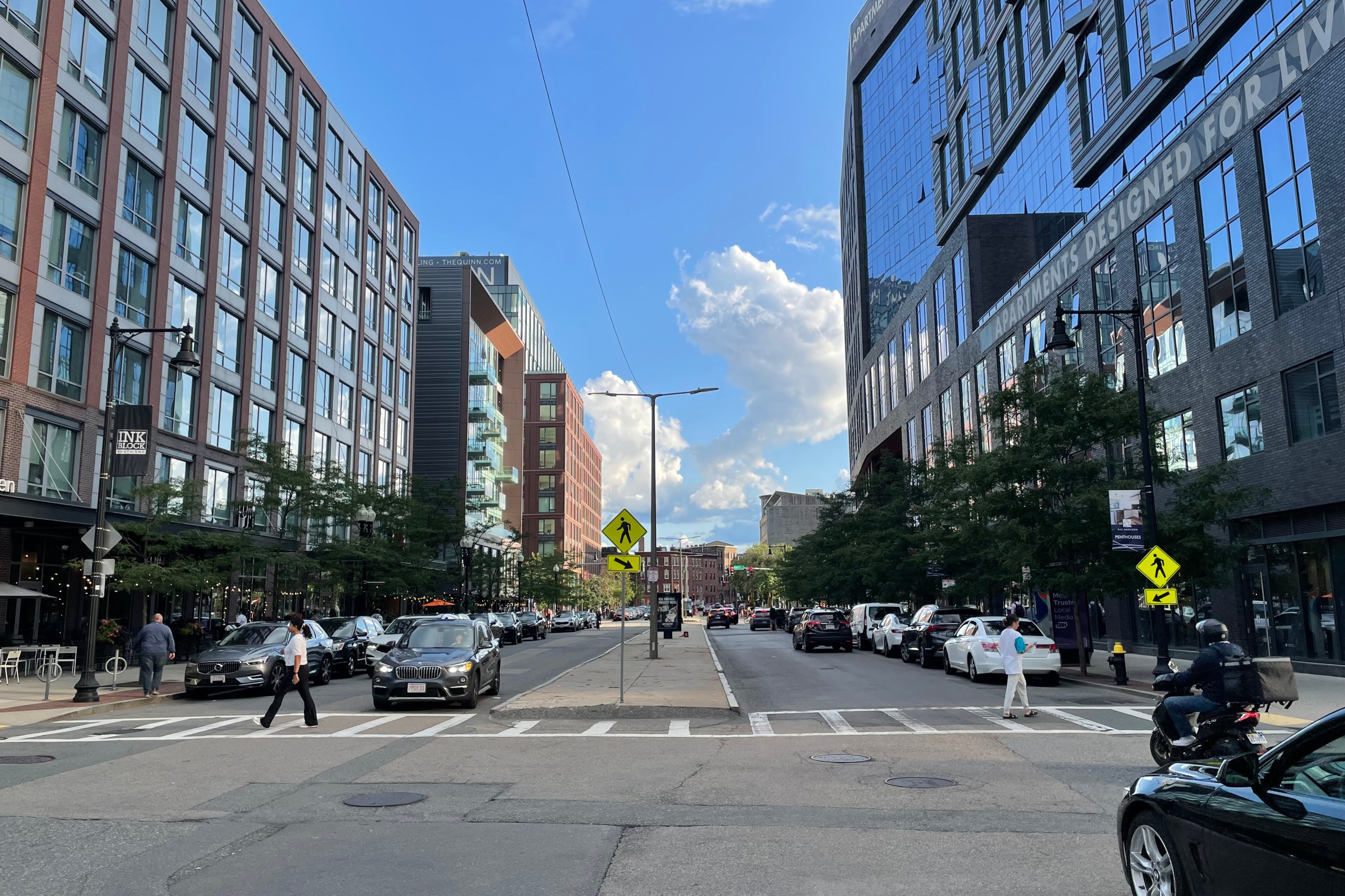 Two pedestrians cross a crosswalk in a wide street with a potholed concrete median. The street is lined with new mid-rise buildings, about 8 to 10 stories tall.