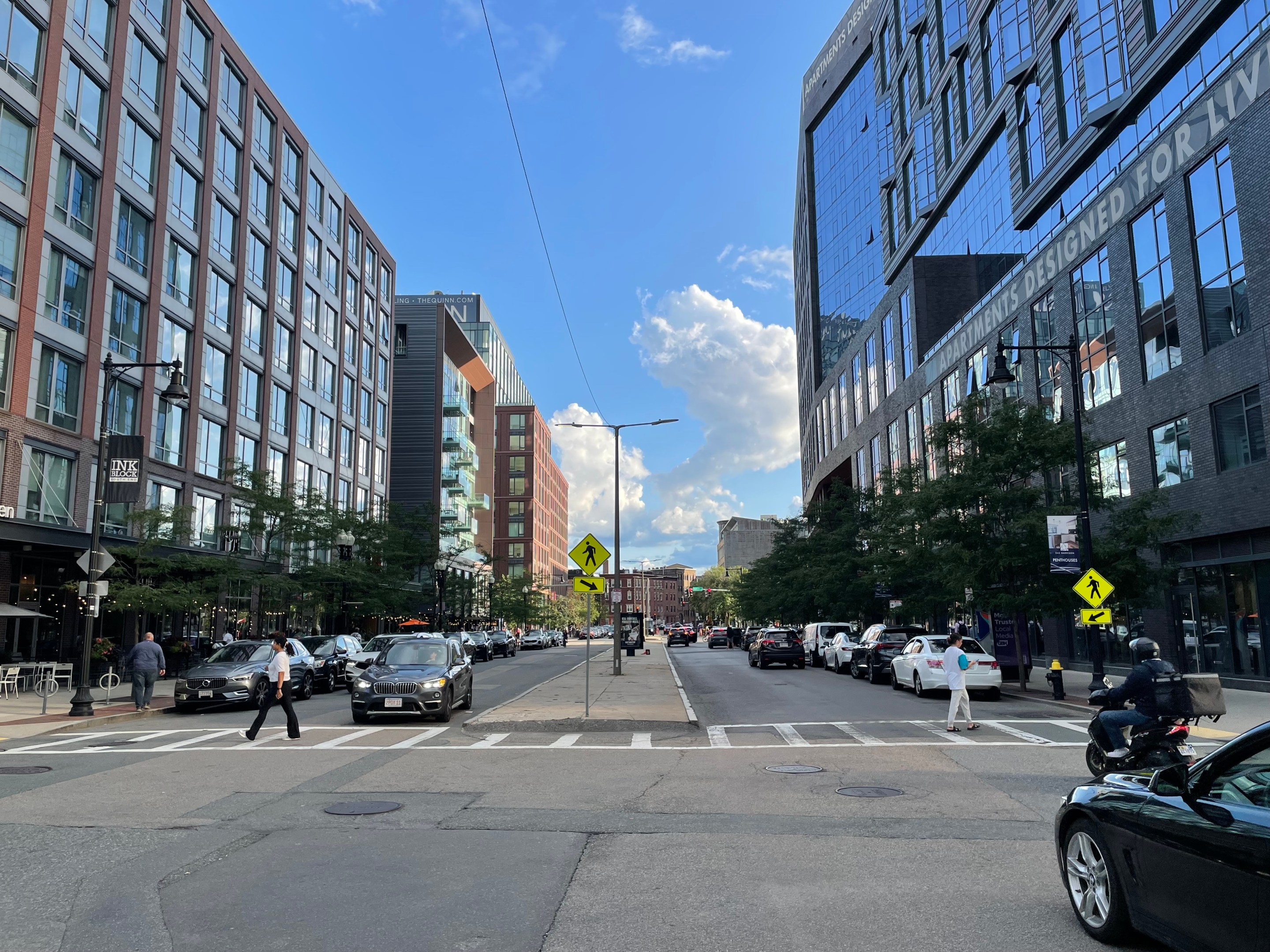 Two pedestrians cross a crosswalk in a wide street with a potholed concrete median. The street is lined with new mid-rise buildings, about 8 to 10 stories tall.