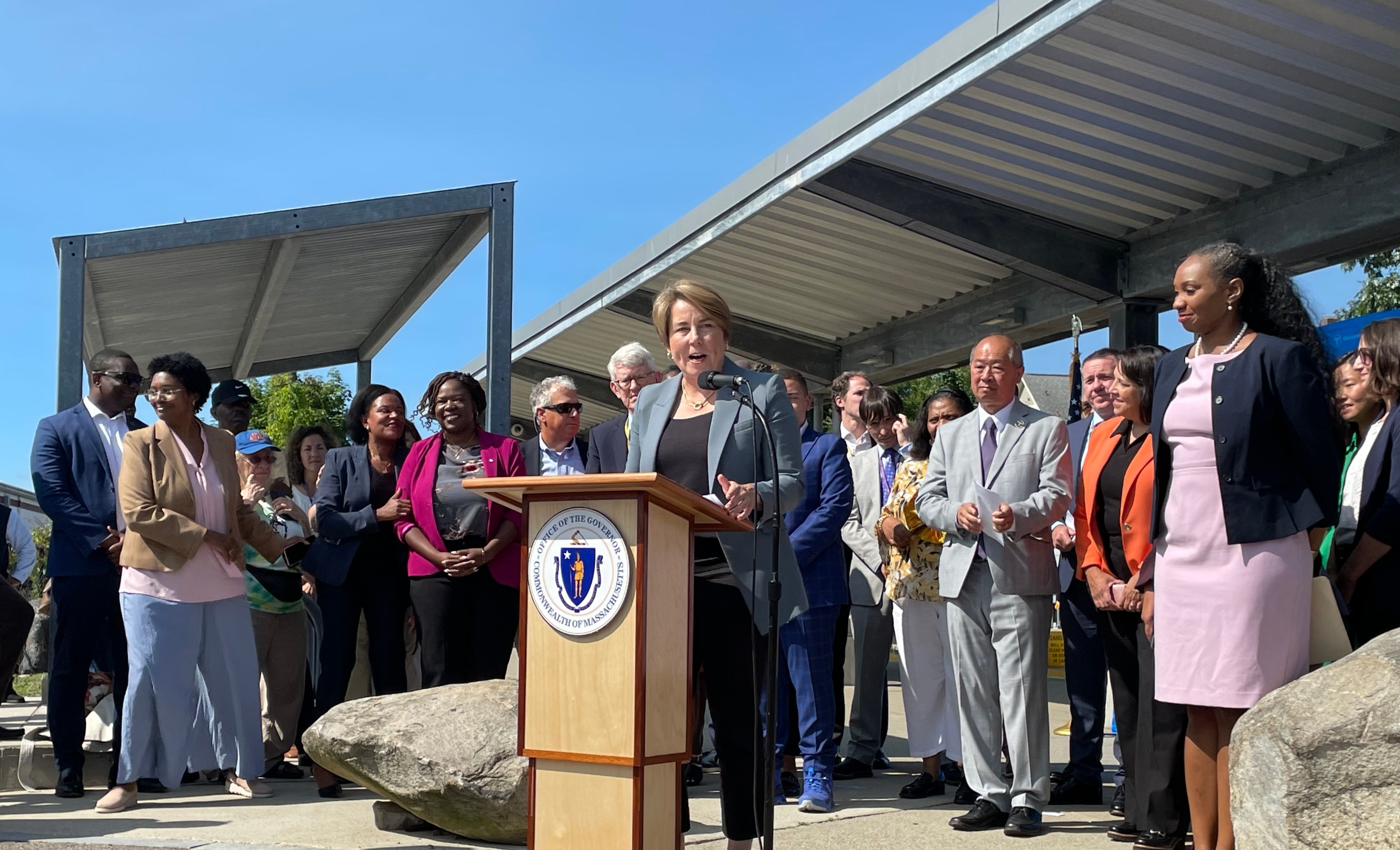 A woman wearing a grey blazer speaks at a podium bearing the seal of the Commonwealth of Massachusetts. Behind her, a crowd of people in business suits, many of whom are smiling, listen. Behind them all is a steel canopy over a commuter rail platform.