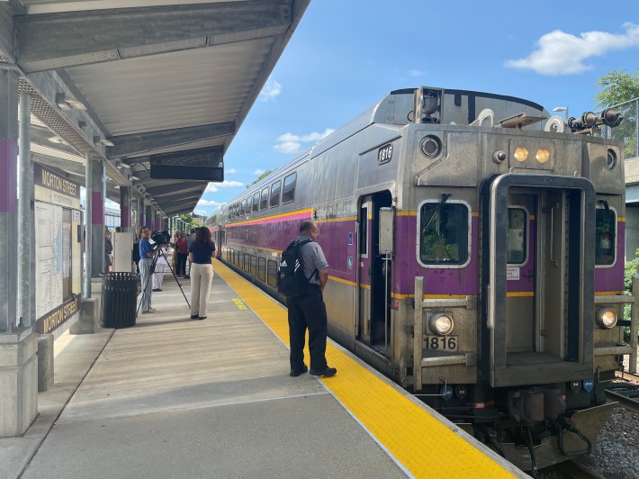 A double-decker commuter rail coach with the MBTA's signature purple stripe waits for a small crowd of passengers to board at a train platform. A sign on the left edge of the photo says "MORTON STREET" above an MBTA commuter rail map.