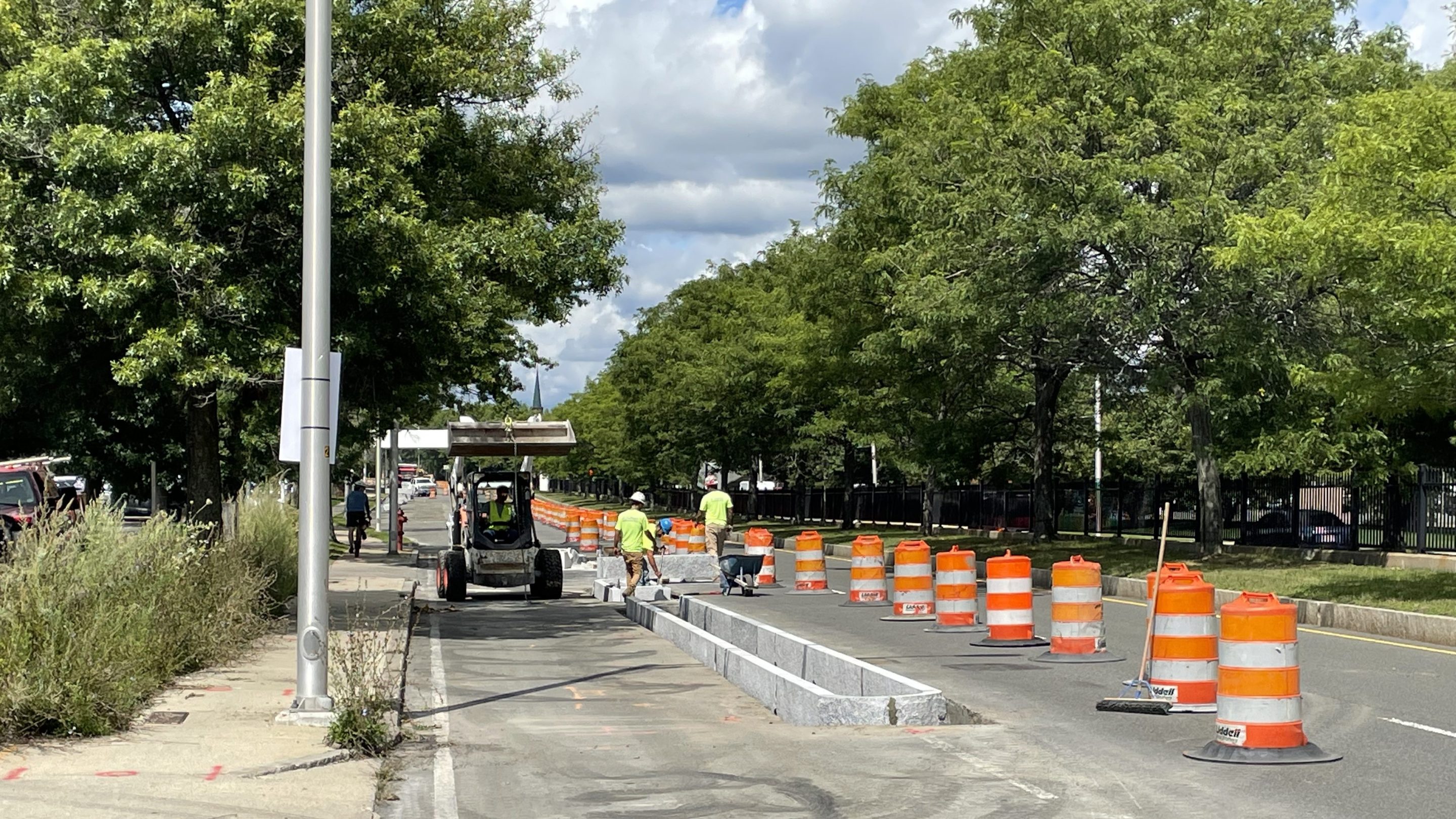 Construction workers place granite curbs into a roadway next to a row of orange construction barrels.