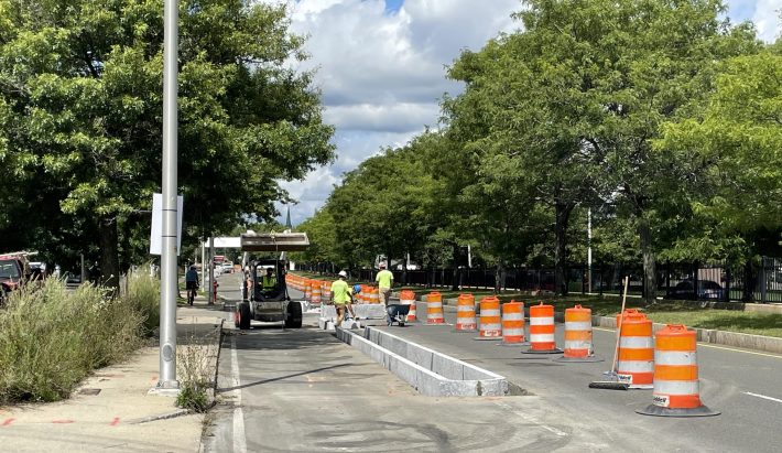 Construction workers place granite curbs into a roadway next to a row of orange construction barrels.