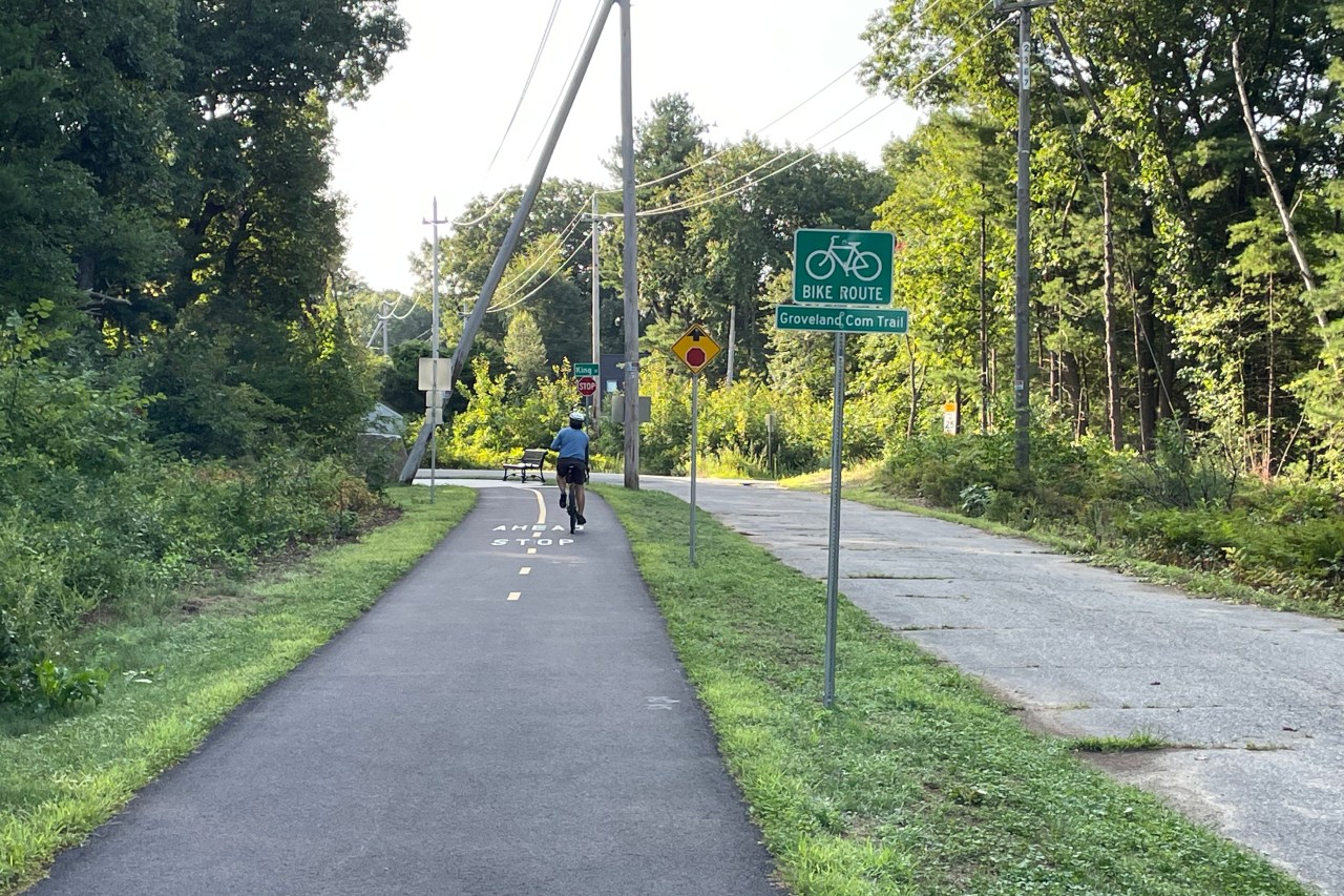 A bike path runs alongside a narrow paved roadway. A green sign reads "BIKE ROUTE: Groveland Com. Trail" and beyond is a stop ahead sign.