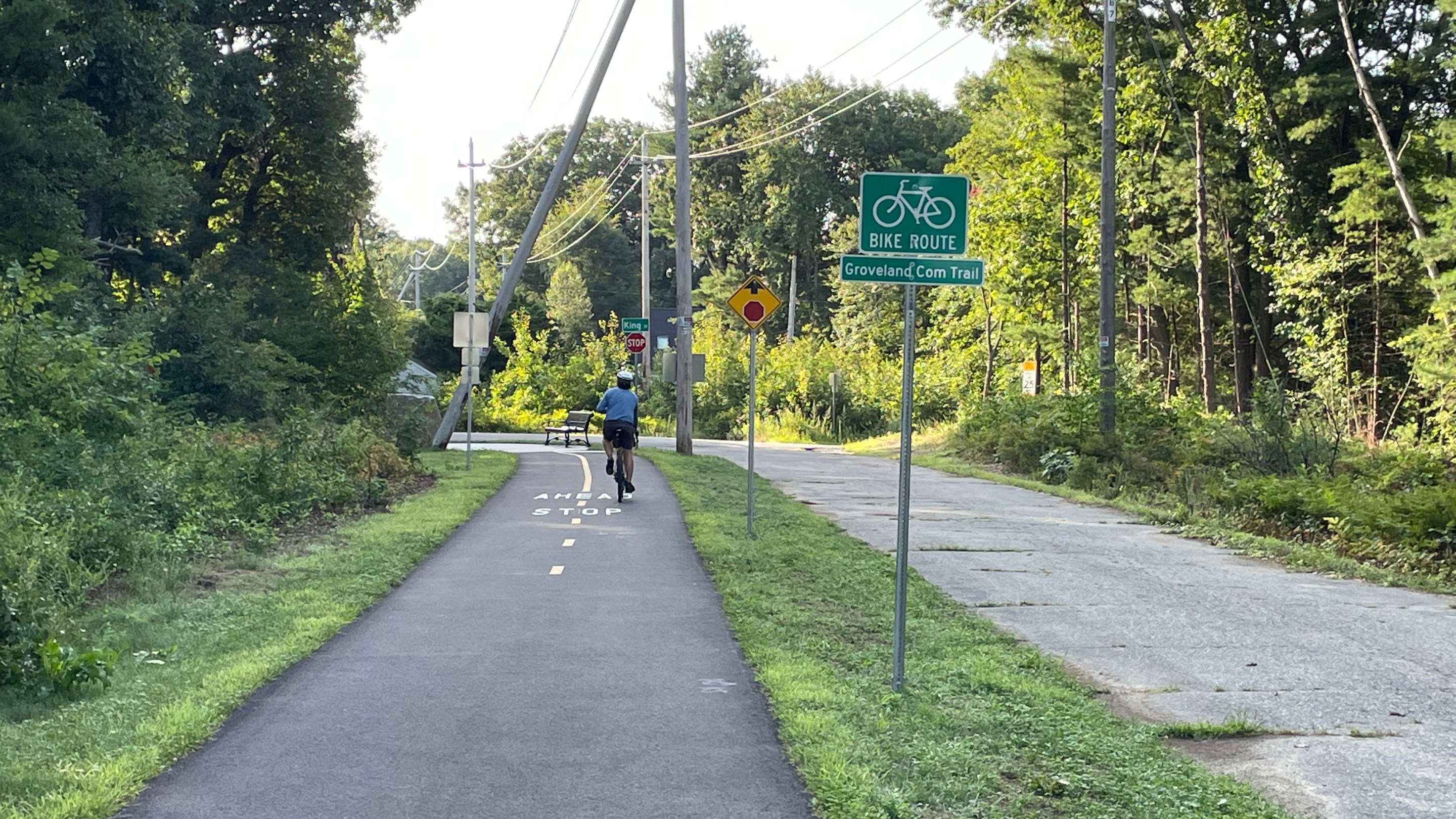 A bike path runs alongside a narrow paved roadway. A green sign reads "BIKE ROUTE: Groveland Com. Trail" and beyond is a stop ahead sign.