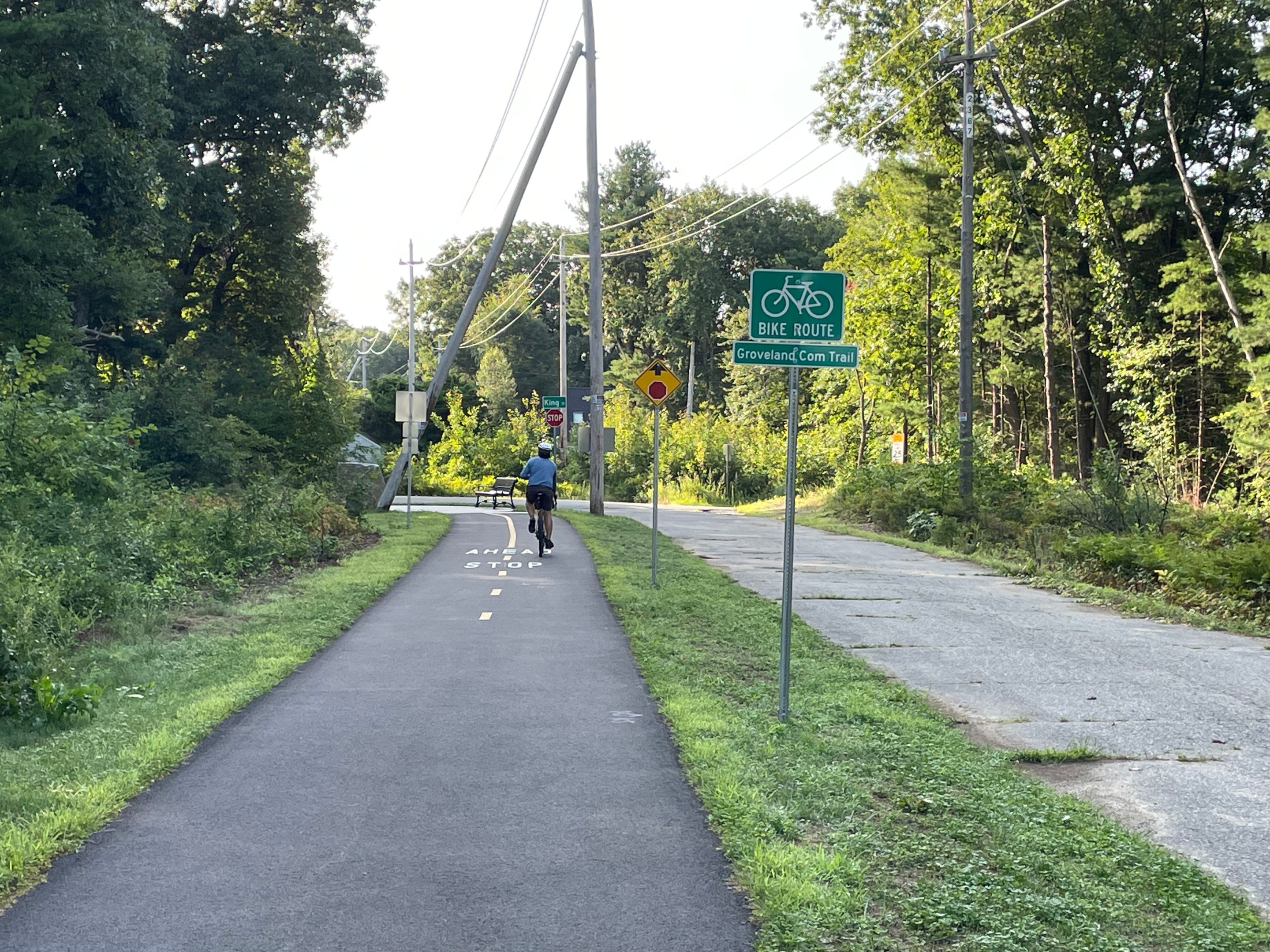 A bike path runs alongside a narrow paved roadway. A green sign reads "BIKE ROUTE: Groveland Com. Trail" and beyond is a stop ahead sign.