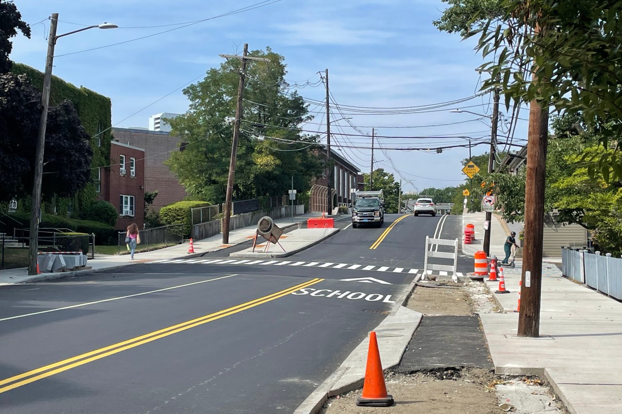 A newly paved street with orange construction barrels and cones along the curb in the foregound, where the sidewalk and bike lane are still under construction.