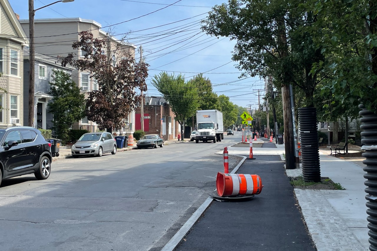 A new sidewalk-level bike lane under construction adjacent to a two-lane city street with a few cars parked along the opposite curb. Cones and construction barrels litter the bike lane which is still awaiting a final layer of pavement.