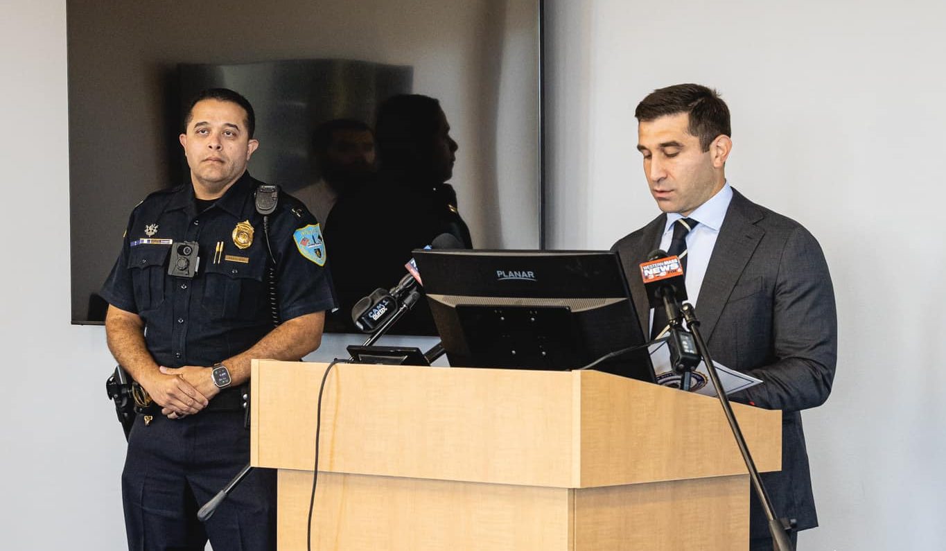 A man in a police uniform stands behind a man in a dark suit reading notes at a podium.