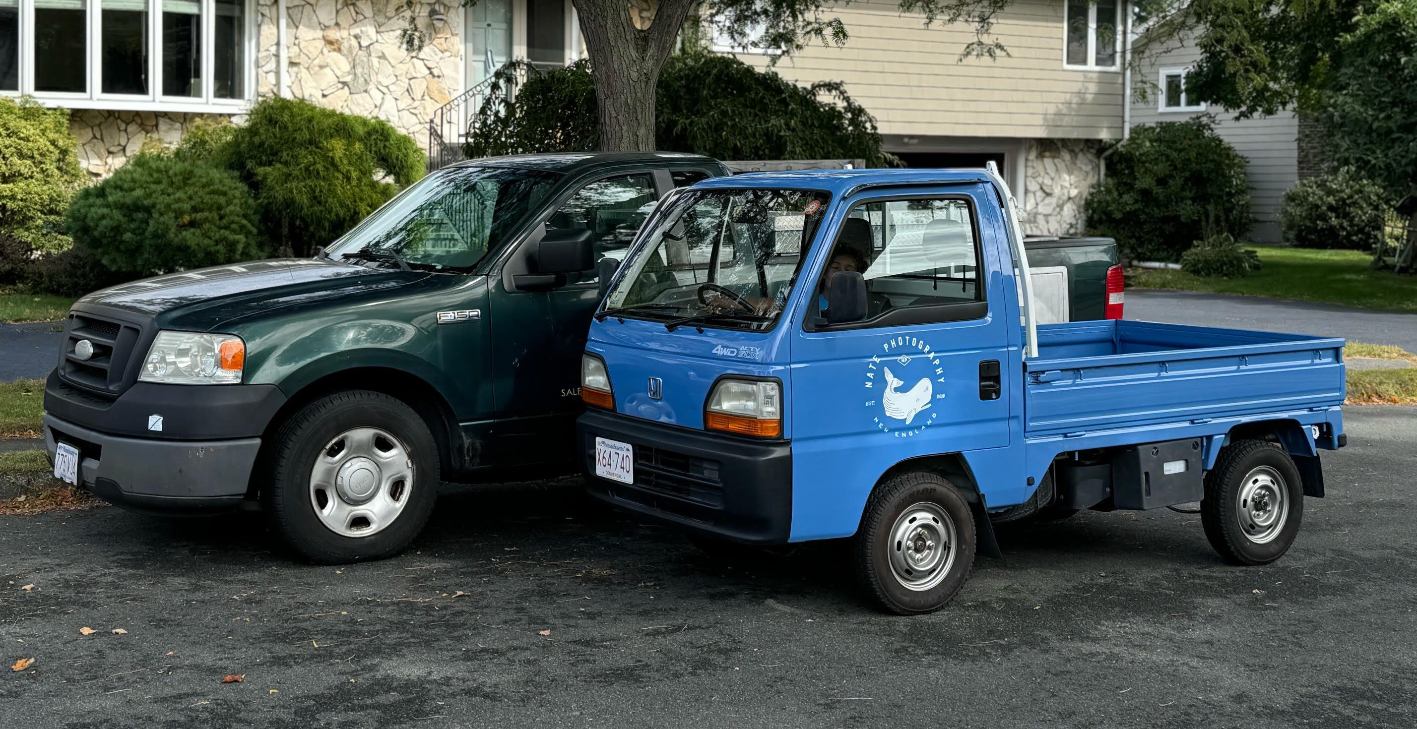 Two trucks parked side-by-side in a suburban neighborhood. In the foreground is a small blue kei truck, with a short two-seater cab and a pickup bed with fold-down side panels. Behind it is parked a much larger green Ford F-150 pickup truck. Both vehicles are roughly the same height but the kei truck is considerably shorter, with a flat front end that's roughly even with the F-150's side mirrors.