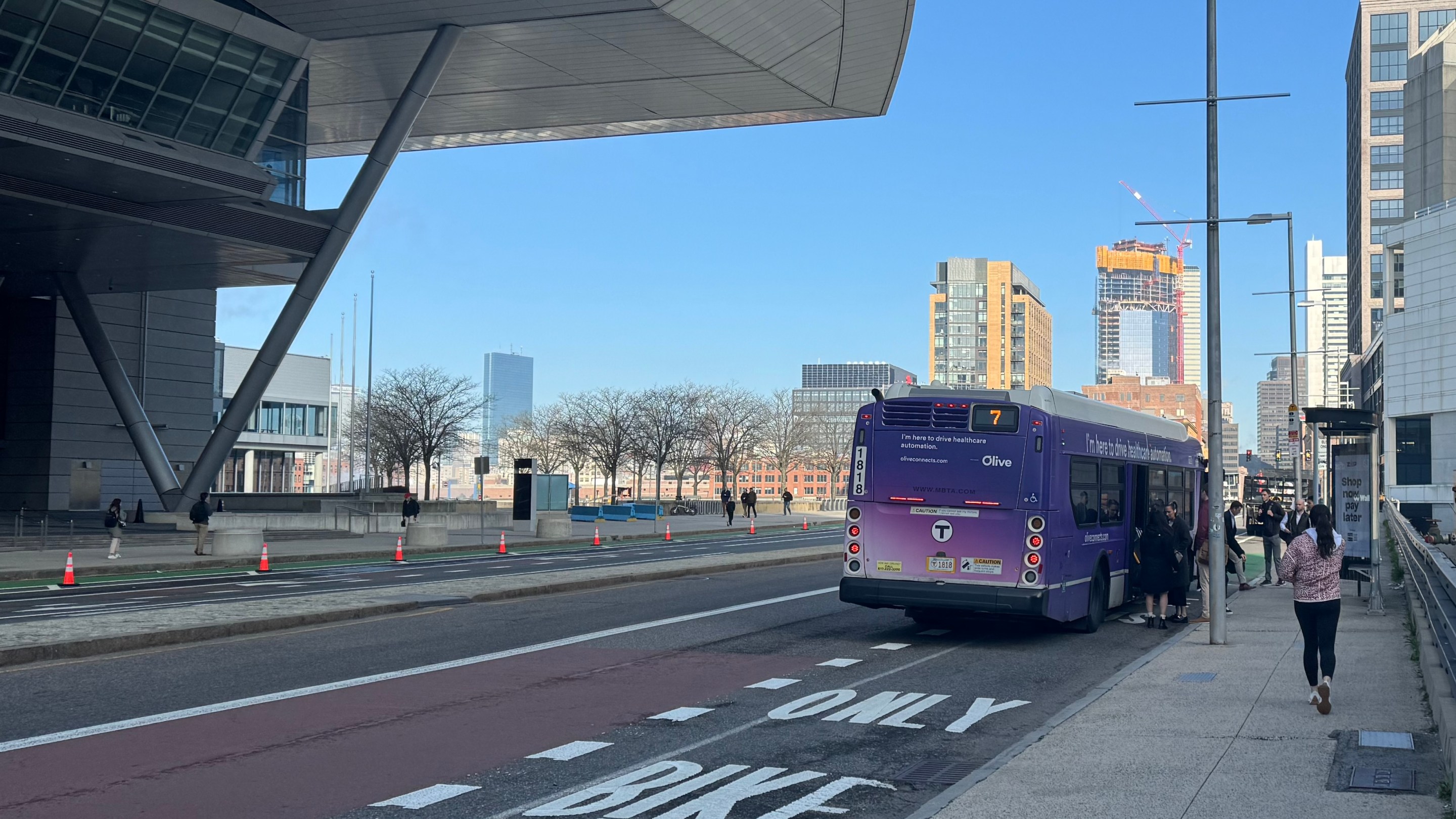 A group of people gather near the doors of an MBTA 7 bus that is pulled over next to a bus shelter on an otherwise-empty multi-lane street with skyscrapers visible on the horizon. On the left side of the photo the massive cantilevered roof of the Boston Convention Center looms overhead.