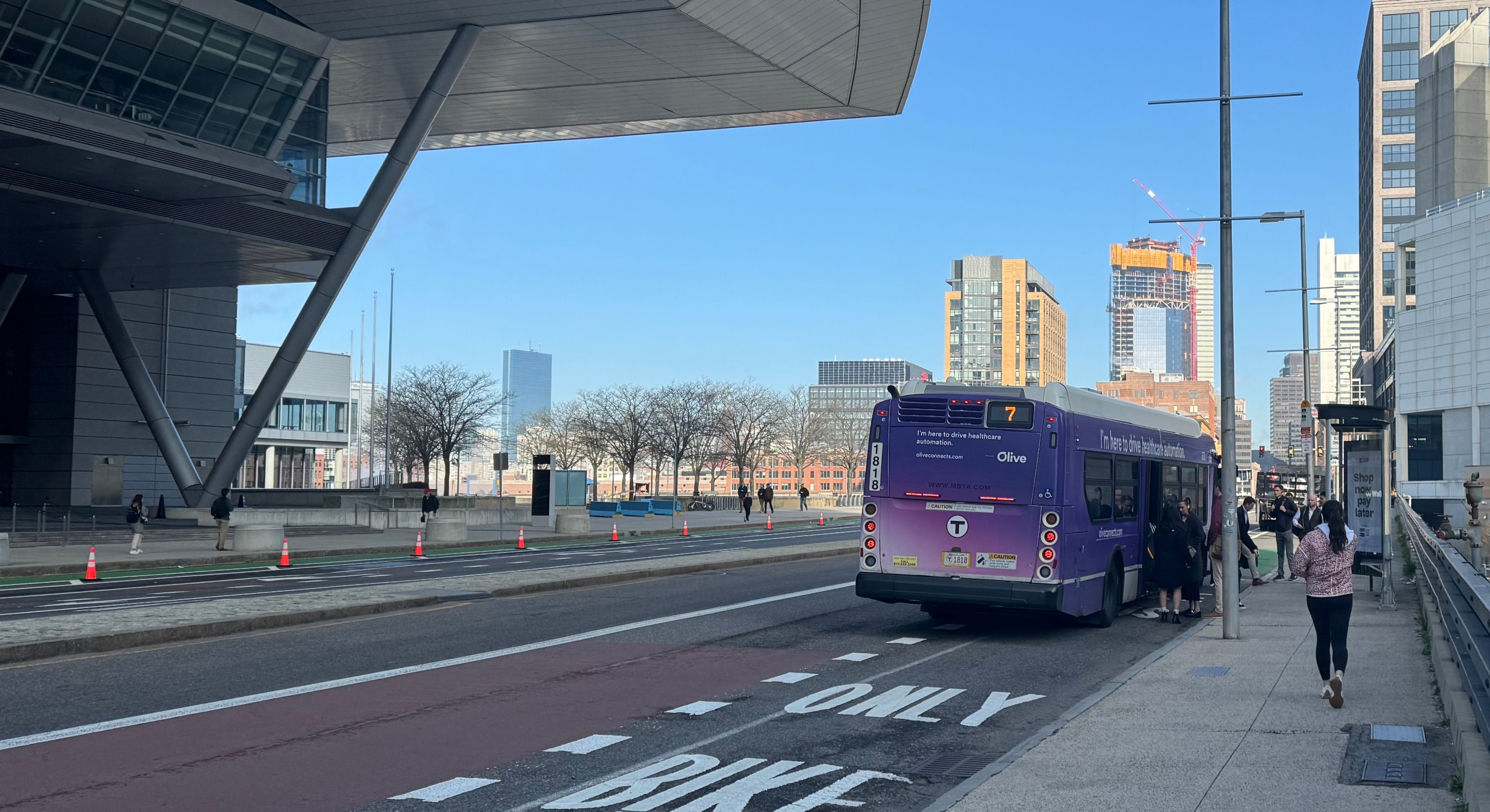 A group of people gather near the doors of an MBTA 7 bus that is pulled over next to a bus shelter on an otherwise-empty multi-lane street with skyscrapers visible on the horizon. On the left side of the photo the massive cantilevered roof of the Boston Convention Center looms overhead.