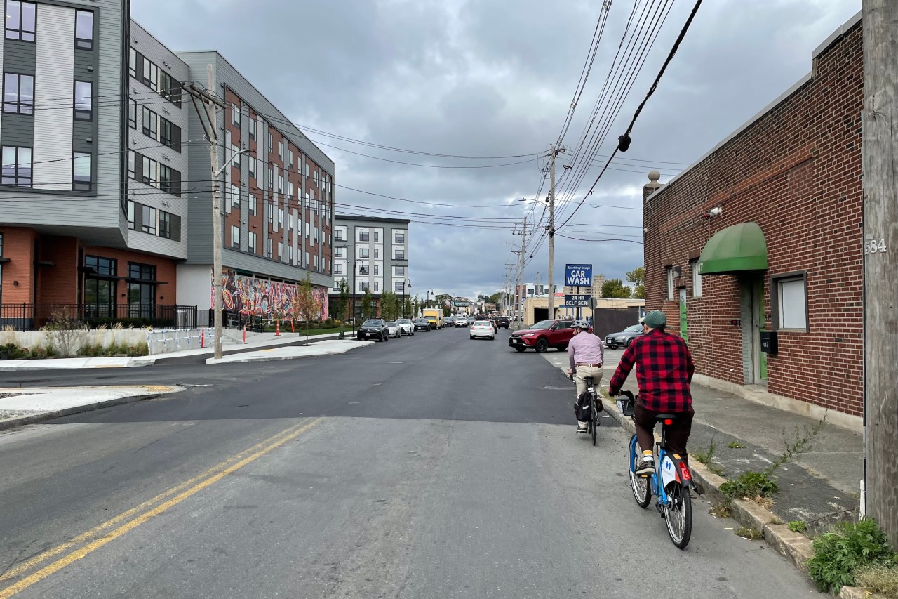 Two people on bikes ride along a city street next to a new 5-story apartment building along the left side of the street. The right side of the street has an old single-story brick warehouse building and power lines above.