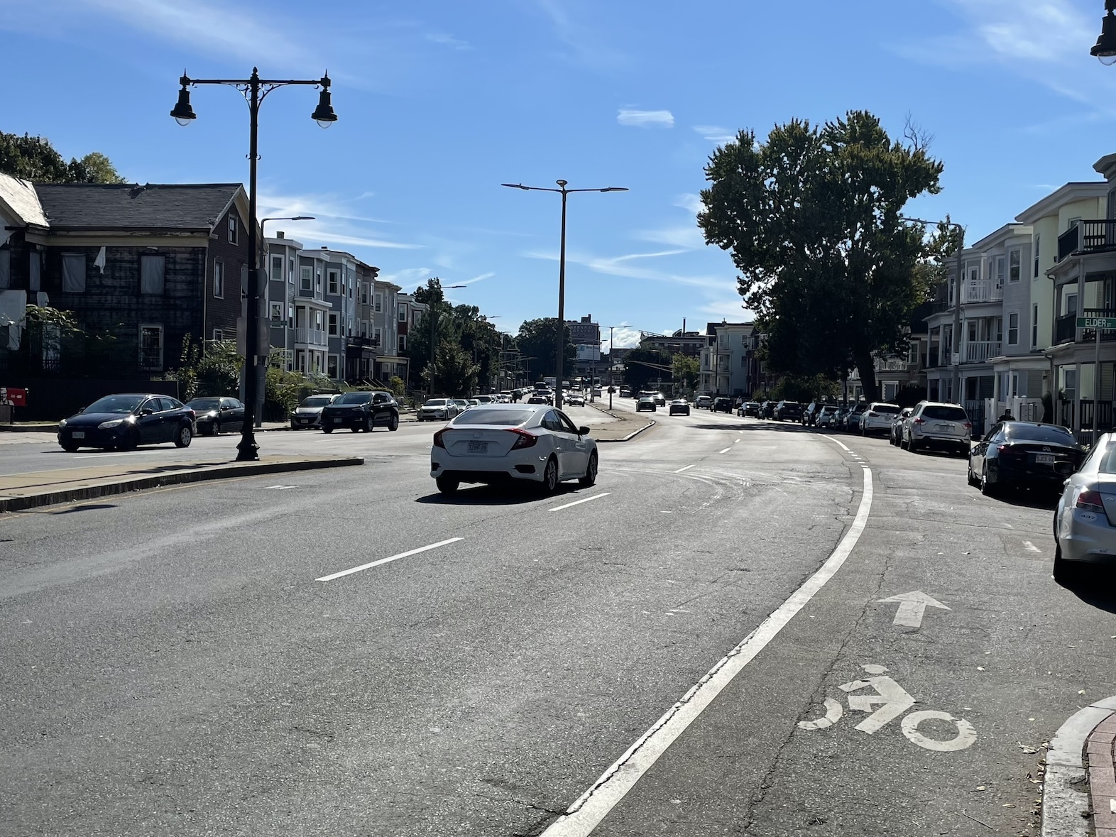 A wide five-lane street with a concrete median in the middle that's lined with tall street lamps. Either side of the broad street is lined with triple-decker apartment houses. Most of the picture is empty asphalt, but there are a few large trees scattered along the street's sidewalks in the distance.