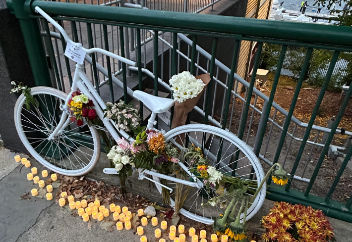 A bicycle painted all white is chained to a fence with bouquets of flowers tied to its frame and a pot of chrysanthemums at its side. The bike's wheels are surrounded by dozens of votive candles.