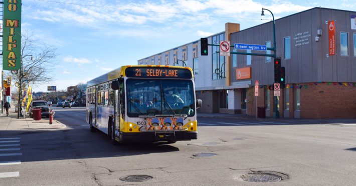 A city bus on route 21, labeled "Selby-Lake," drives down Bloomington Avenue in Minneapolis. Surrounding businesses include a green sign for "Los Milpas" and "The Family Partnership" building on the right. The scene shows a clear, sunny day with pedestrians and parked cars.