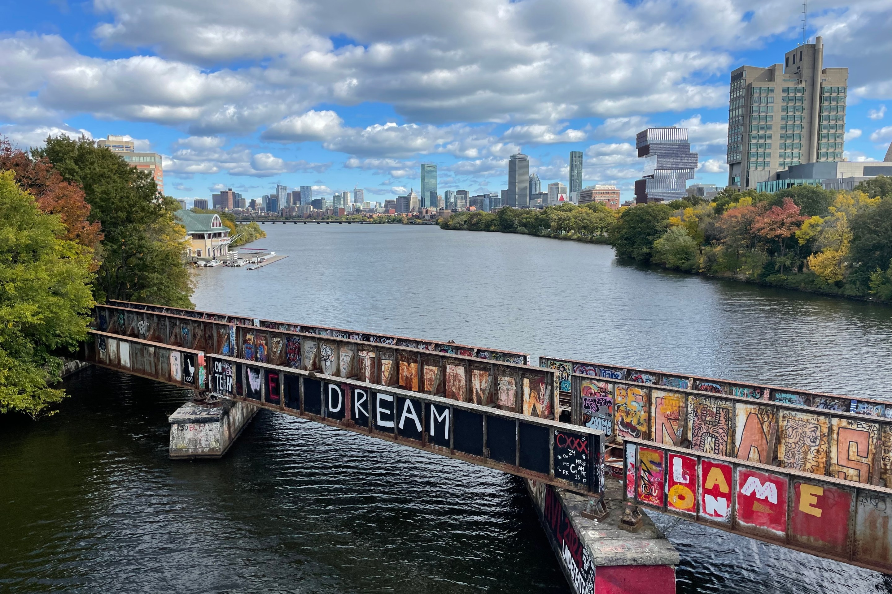 A rusted bridge covered with graffiti spans a river with a city skyline in the distance.