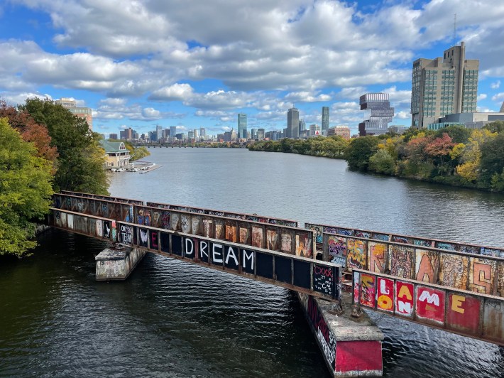 A rusted bridge covered with graffiti spans a river with a city skyline in the distance.