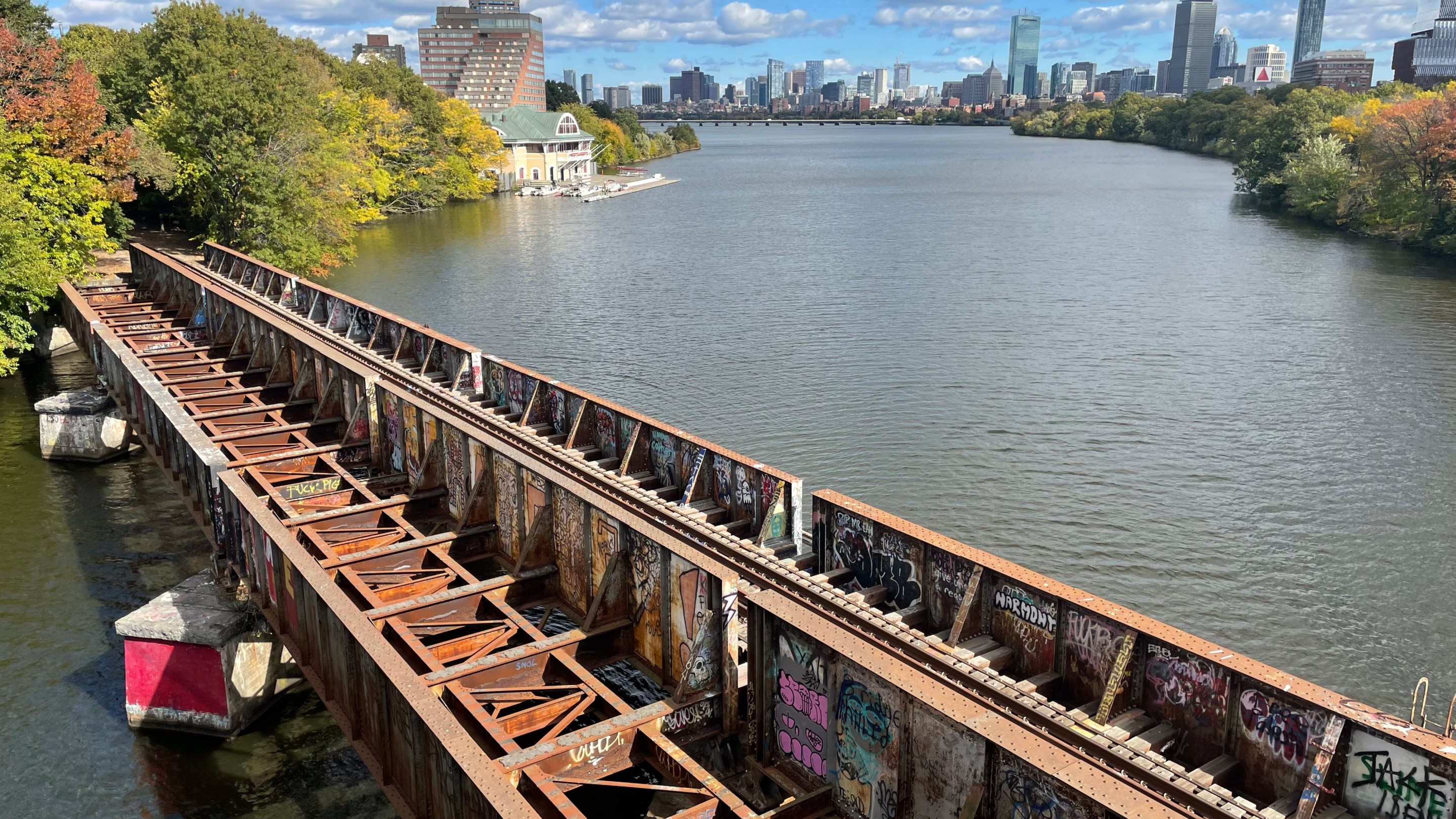 A rusted bridge covered with graffiti spans a river with a city skyline in the distance.