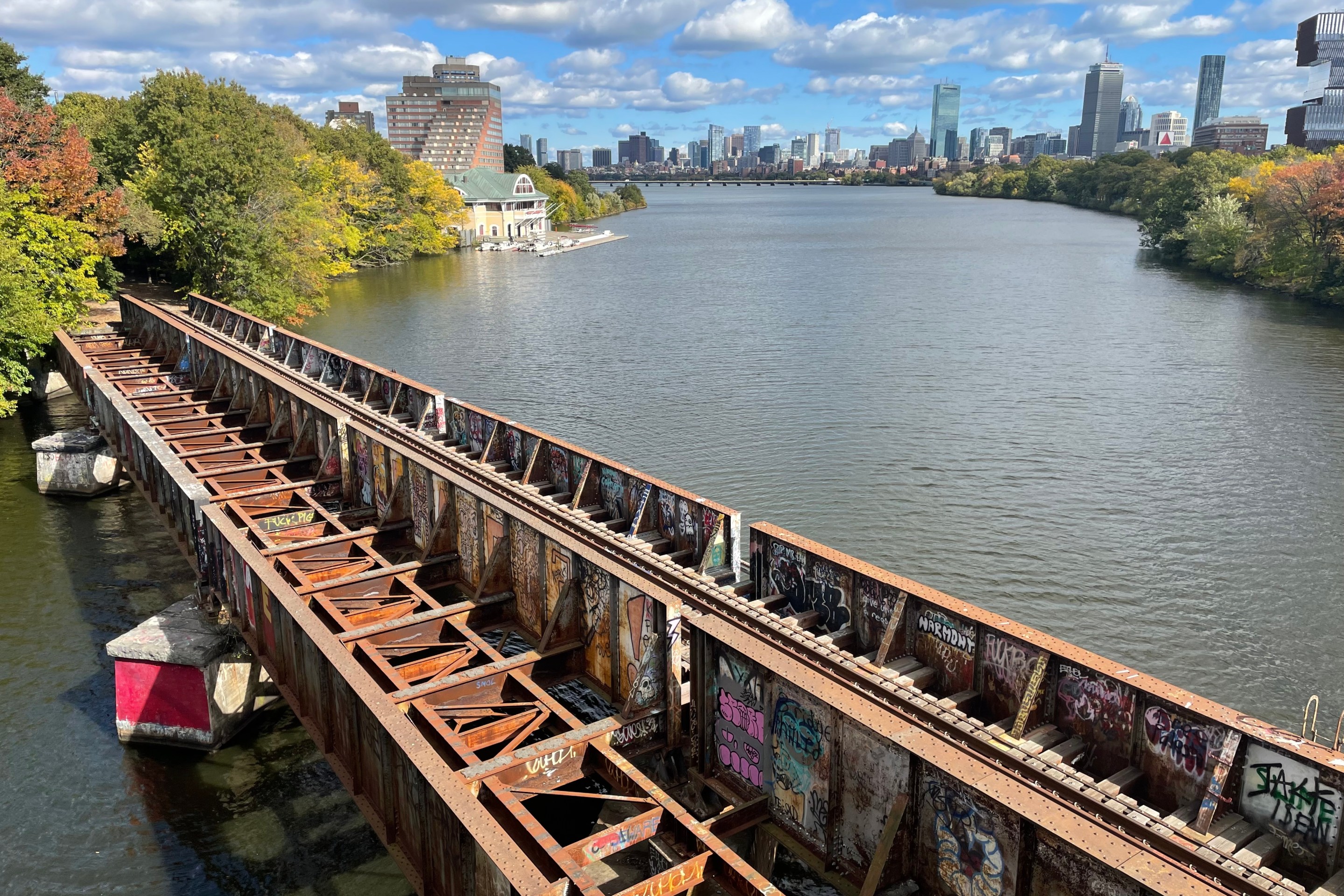 A rusted bridge covered with graffiti spans a river with a city skyline in the distance.