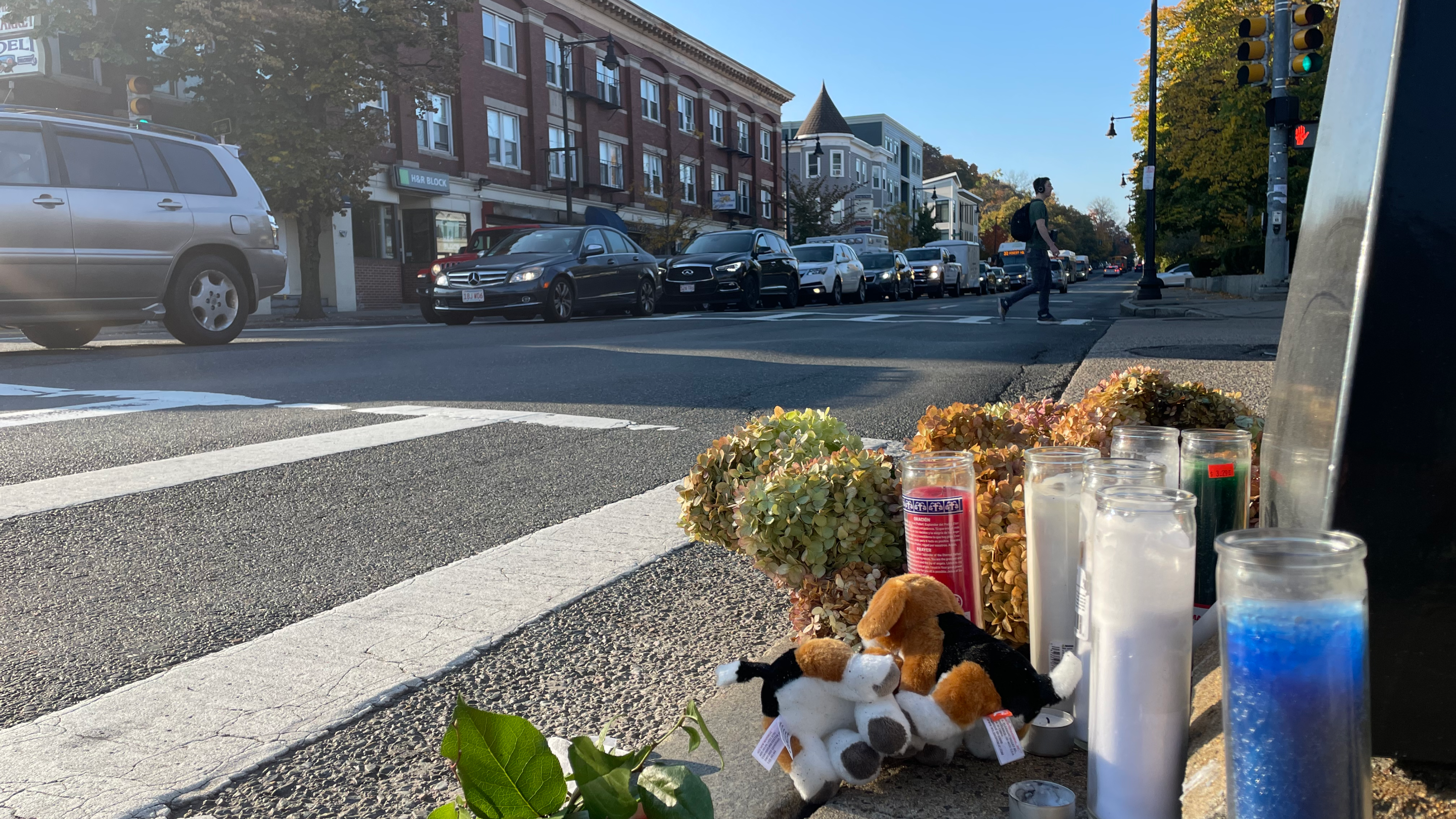 Votive candles and flowers lie on the curb next to a crosswalk on a wide four-lane street.