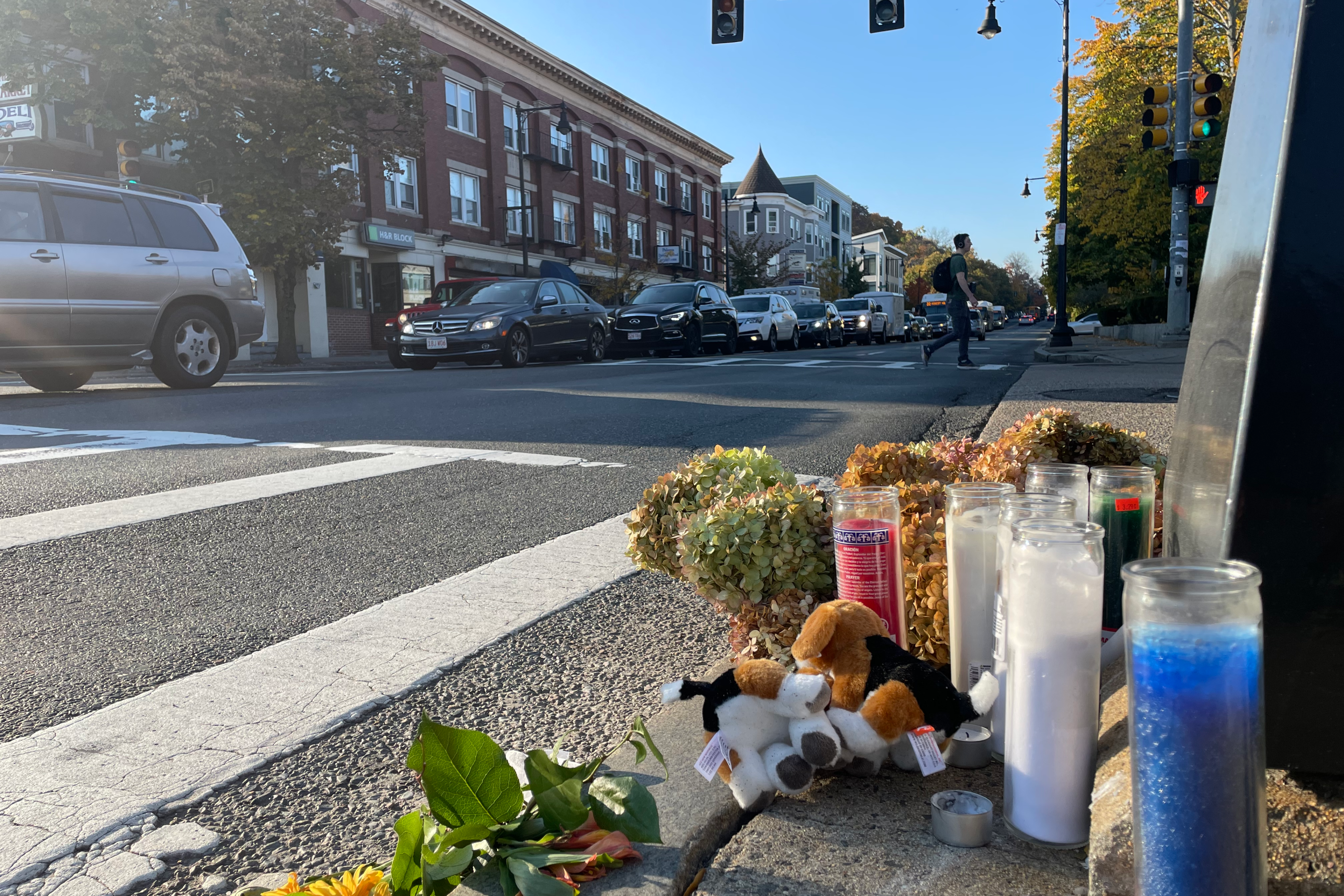 Votive candles and flowers lie on the curb next to a crosswalk on a wide four-lane street.