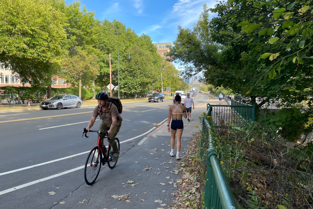 Several pedestrians and a person on a bike pass each other on a narrow sidewalk next to a wide multi-lane highway.