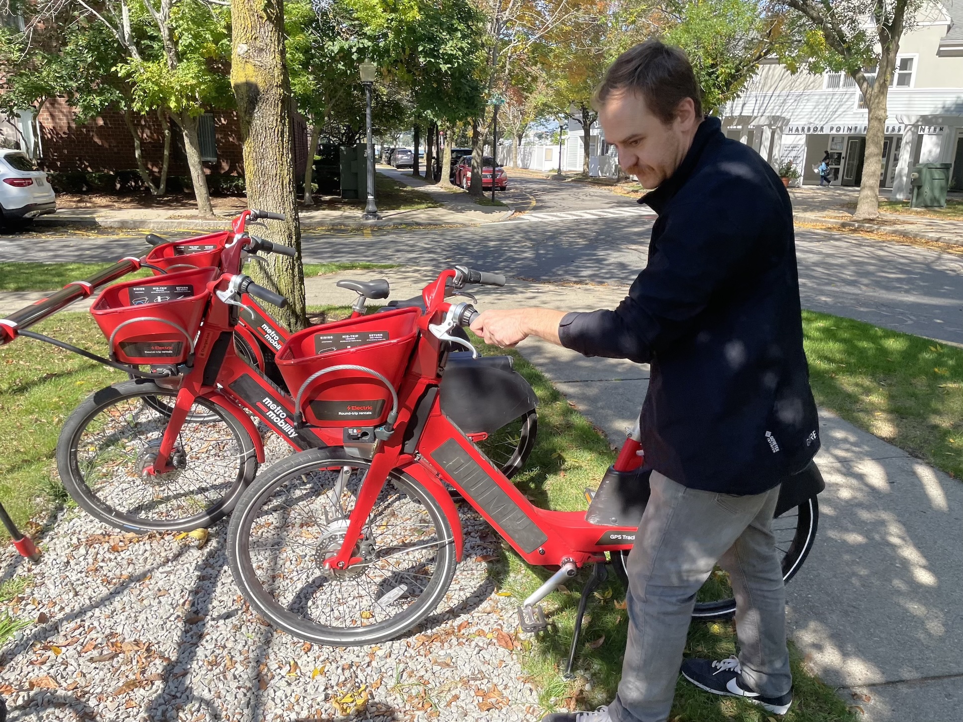 A man wearing a black coat walks a heavy red e-bike with a large front basket into a bike rack next to a sidewalk in a leafy residential neighborhood