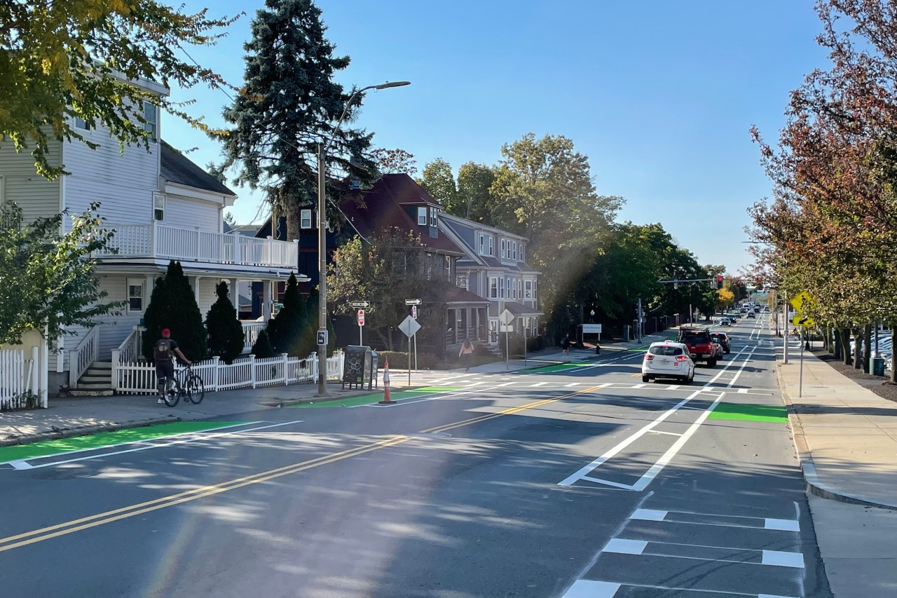 A city street under a sunny blue sky. The street features two bike lanes along each curb on either side of the street with hatched lines to buffer the bike lane from moving car traffic in the middle of the street.