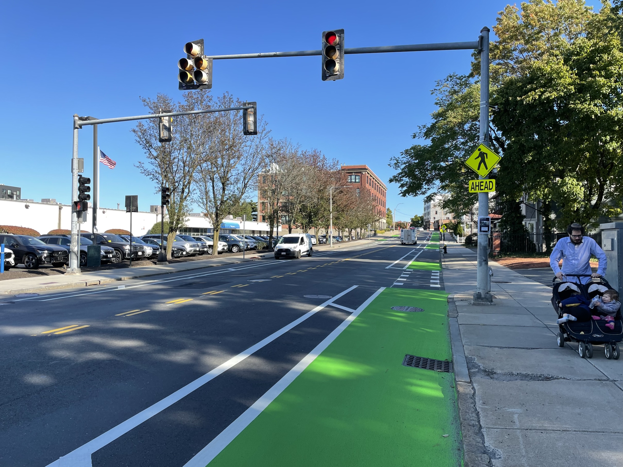 A city street and traffic signal. The street features two bike lanes along each curb on either side of the street with hatched lines to buffer the bike lane from moving car traffic in the middle of the street. A man pushes a stroller with two children on the sidewalk next to the street.