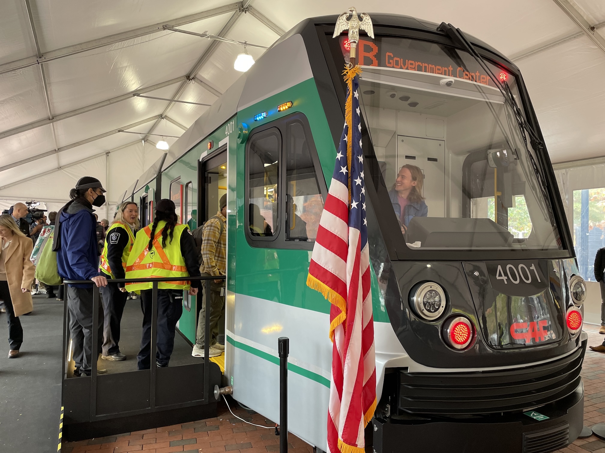 A full-size mockup of a Green Line train sits behind an American flag under a white tent. On a ramp to the left, people wait to enter the doors of the mockup.