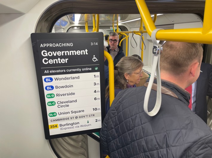 A mockup of a passenger information screen inside a light rail vehicle. The display reads "Approaching Government Center" next to a wheelchair icon. Below that, the screen reads "All elevators currently online" followed by a list of transit connections and estimated arrival times for station connections to the Blue Line, other Green Line branches, and the 354 bus.