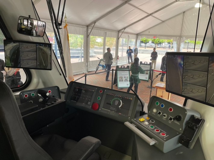 An operator's console at the front of a train car, with various buttons and screens in front of a chair, and a wide front window.