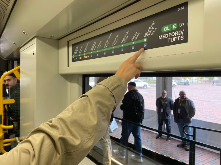 Someone points to a strip map showing part of the MBTA Green Line on a screen above the window of a train car.