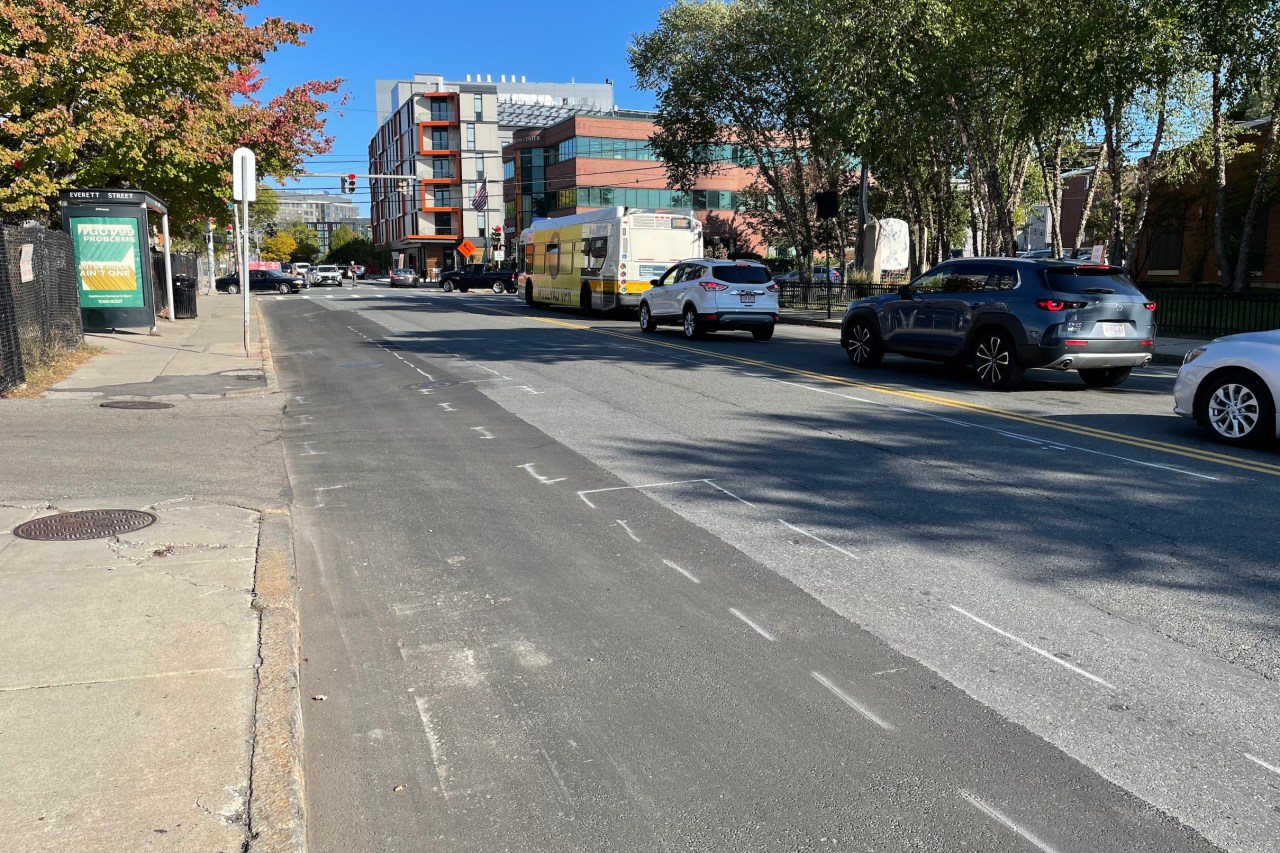 A city street and traffic signal in the distance with a row of cars waiting for the light. Spray-painted markings outline a future bike lane in the foreground.