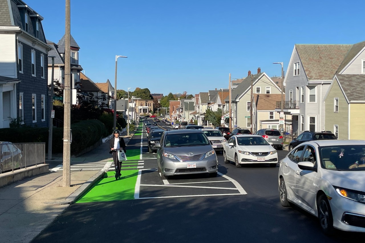 A recently-paved street lined with 2 and 3-story houses with gable roofs. The street features a bike lane on the left, with a man riding a scooter towards the camera. Between the bike lane and moving traffic in the center of the street is a row of parked cars.