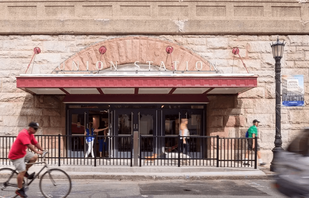 Several people walk by the entrance to an old stone building with a broad stone arch and a cantilevered metal canopy over the doors. In the foreground a middle-aged man rides a bike on the street.