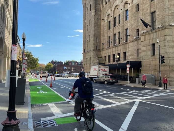 A person on a bike rides on a green-painted bike lane on the edge of a multi-lane city street next to a 19th century masonry building