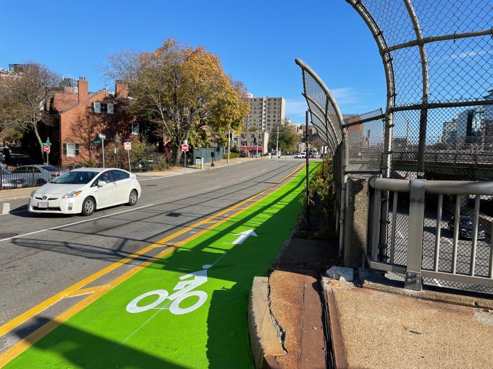 A green bike lane runs alongside the edge of a multi-lane street above a depressed highway. Older brick houses line the street on the other side away from the highway on the left edge of the photo.